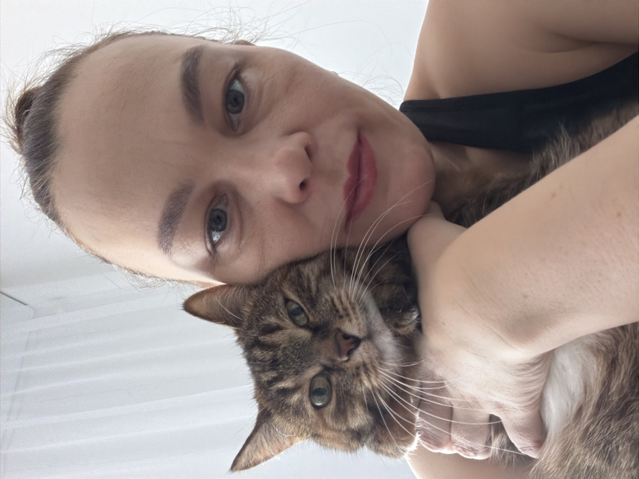 The image displays a close-up, upward-angled shot of an adult female subject holding a tabby cat. The female has light skin, blue-grey eyes, and her hair is pulled back into a bun, revealing a high forehead. She wears red lipstick and a black top, with a strap visible on her right shoulder. Her left hand is visible supporting the cat's body, bringing the cat's head to rest against her left cheek and chin. Both the female subject and the cat are looking directly towards the camera. The cat is brown with darker tabby stripes and has greenish-yellow eyes and prominent white whiskers. The background is uniformly light, with a sheer white curtain or fabric visible on the upper left, indicating an indoor setting with natural light. No specific identifying features of the city of Chaikovsky, Russia, are discernible in the visual content.
