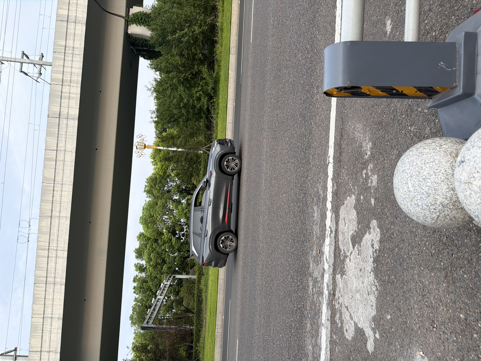 A grey SUV with red trim is driving on a paved road. The car is in the center of the image, moving from left to right. In the background, there is a large concrete overpass, green trees, and utility poles with wires. The foreground shows a portion of the road with white lane markings, a white barrier with yellow and black reflective tape, and two round, stone-like bollards. The sky is overcast. This scene is likely located on a highway or major road in the city of Jiangjin, China, given the presence of the overpass and the general urban/suburban landscape.