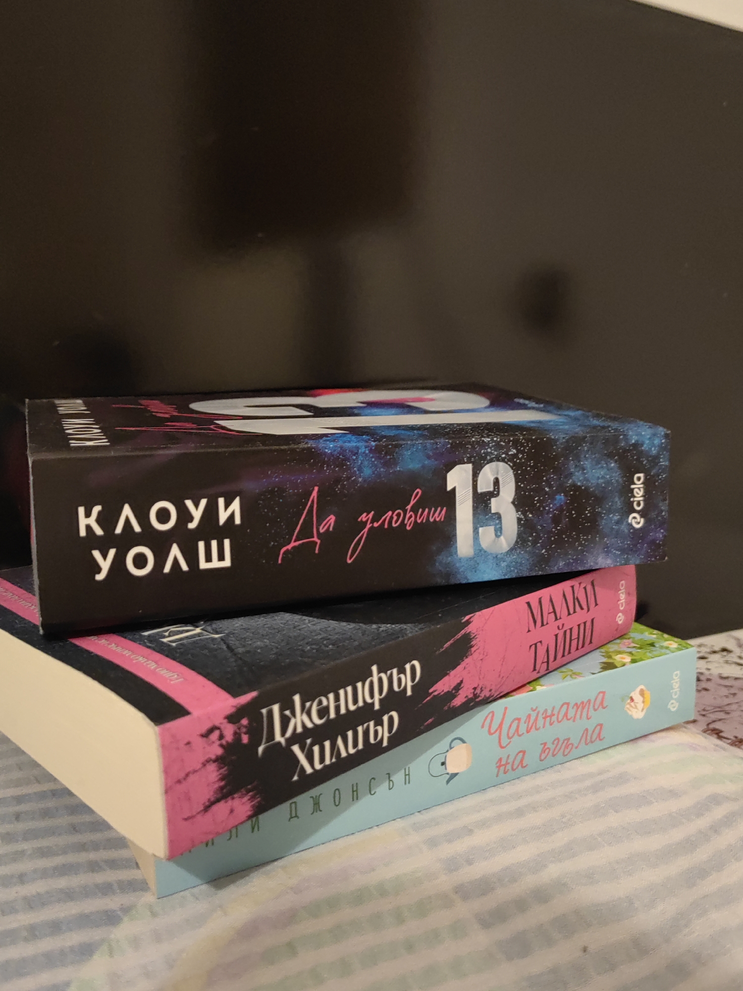 The image shows a stack of three books on a bed. The top book has a black cover with a cosmic design and text in white and pink. The text on the cover reads "КЛОУИ УОЛШ" (Kloui Wolsh) and "Да уловиш 13" (To Catch 13). The middle book has a pink cover with black text that appears to be "МАЛКИ ТАЙНИ" (Malki Tayni). The bottom book has a light blue cover with text in pink and white, which reads "Чайката на въгла" (Chaykata na Vughla) and "ДЖОНСЪН" (Johnson). The books are stacked horizontally, with the top book slightly angled. The background is dark and out of focus. There is no discernible location in the city of Djerovo, Bulgaria, or any indication of people, actions, or interactions.
