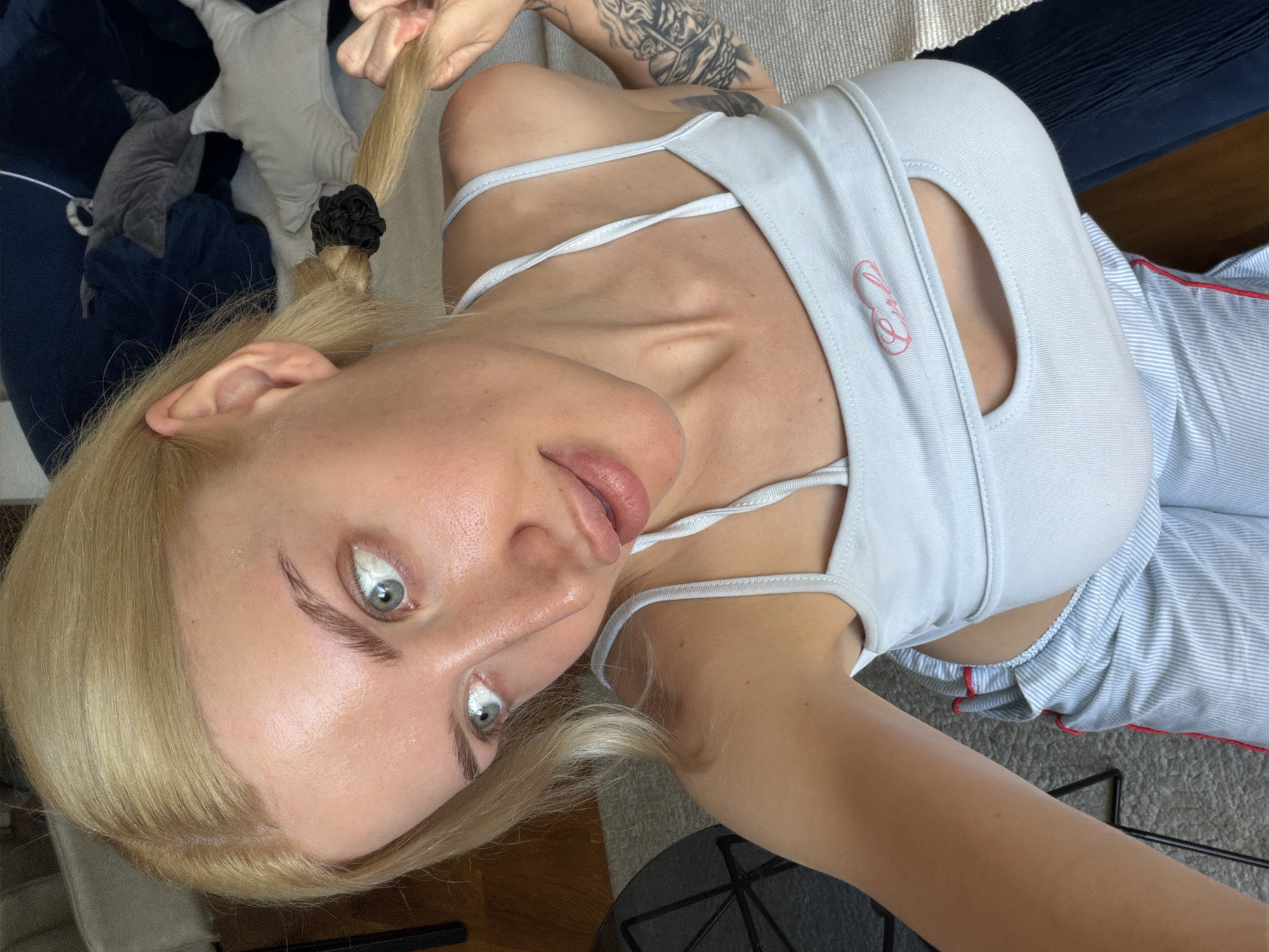 A woman is lying on a couch, looking directly at the camera. She has blond hair, styled in a braid with a black hair tie. She is wearing a light blue top with thin straps and a cutout. Her outfit includes striped pants with a red trim. Her left arm is tattooed. There are pillows and a gray stuffed animal on the couch. The scene is indoors, but the exact location in Belgrade, Serbia cannot be determined from the image.