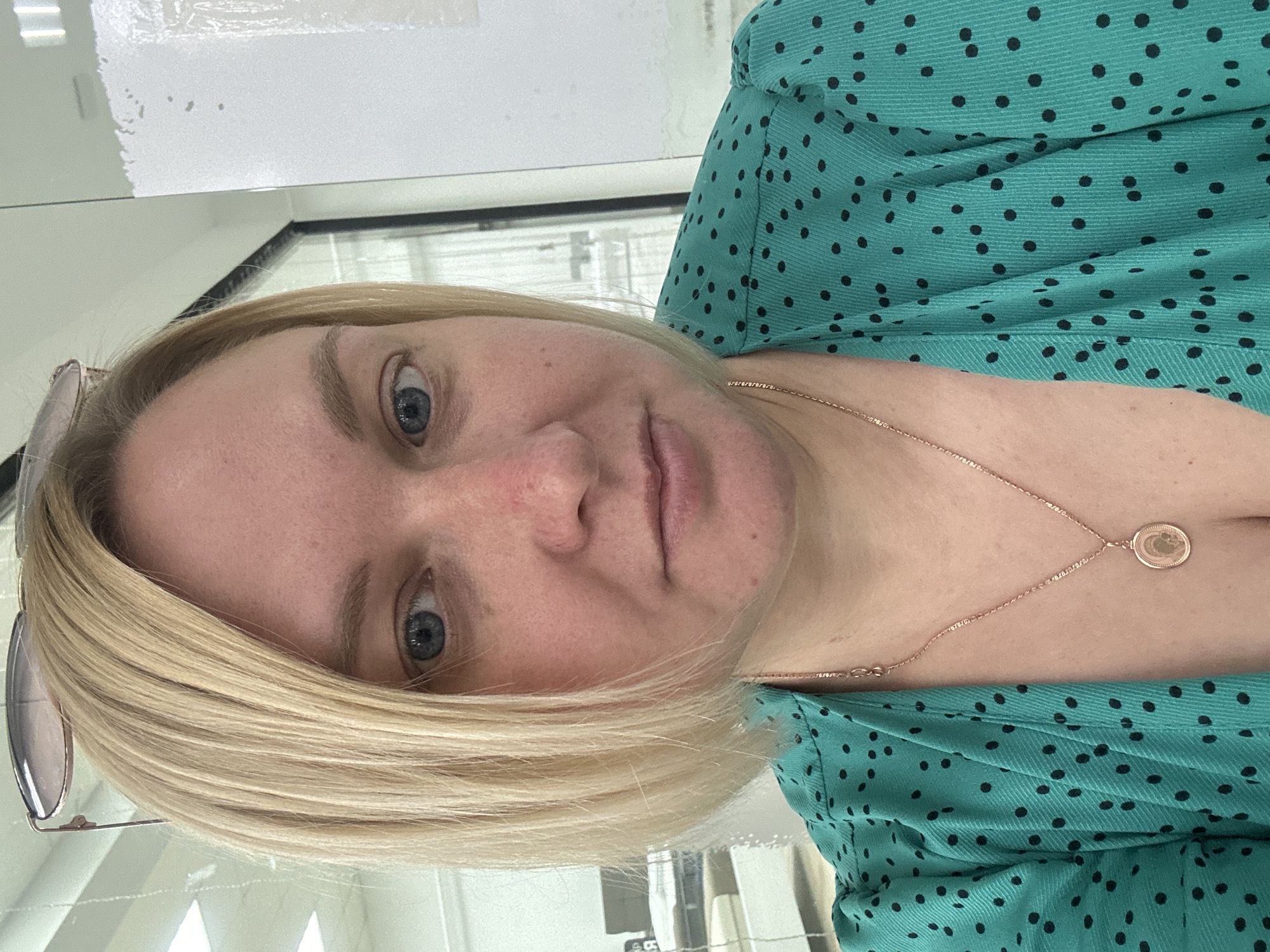A woman with blonde hair and blue eyes is shown in a close-up shot. She is wearing a teal, polka-dotted dress and a rose gold necklace with a circular pendant. Her glasses are perched on top of her head. The background appears to be an office or indoor space with office furniture and fluorescent lighting. The image was taken in Chaikovsky, Russia.