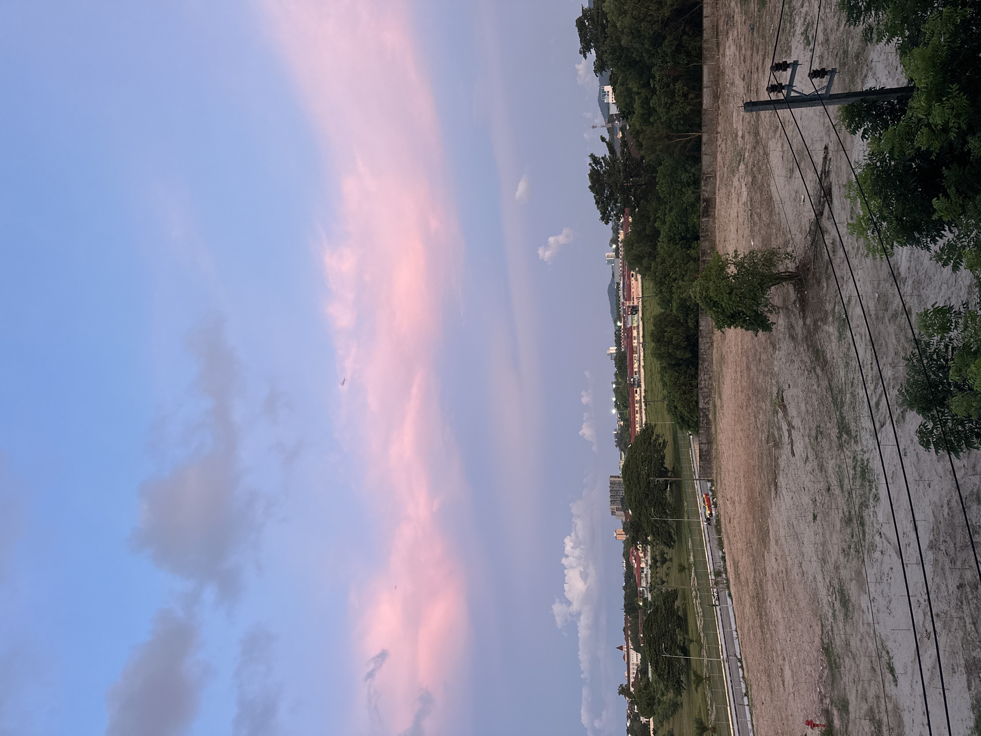 The image depicts a landscape view of a city, likely in the late afternoon or early evening, given the soft pink and blue hues in the sky. The foreground is dominated by a large, bare, sandy lot with a few sparse patches of dry grass and some scattered branches. A solitary, small tree stands in the middle of the lot. Towards the right side of the foreground, a wooden utility pole with multiple insulators and overhead wires is visible.
In the middle ground, a road runs horizontally, bordered by a fence. Two vehicles, one red and one yellow, are on the road. Beyond the road, there is a stretch of green grass, followed by a line of trees and then a collection of buildings. These buildings appear to be residential or institutional, with red-tiled roofs and some taller structures in the distance. The background shows a slightly hazy horizon with more buildings and a range of hills or mountains.
The sky is a prominent feature, transitioning from a light blue at the top to a soft pink and lavender near the horizon. There are scattered clouds, some tinged with pink from the setting sun. The overall impression is one of a developing urban area with natural elements and a picturesque sky. The image does not contain any discernible people or specific actions, but rather captures a static view of the environment.