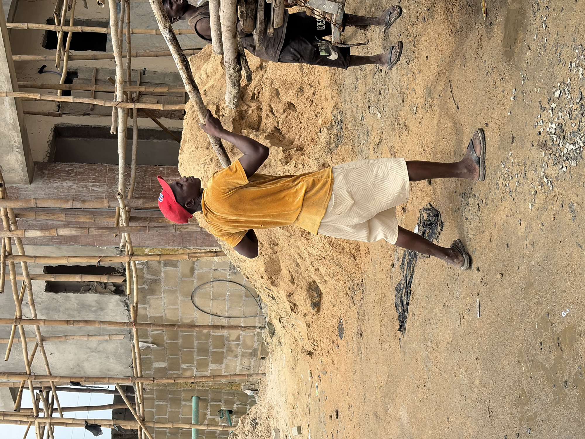 In Igboefon, Nigeria, a construction scene unfolds with two individuals engaged in labor. The foreground features a man wearing a yellow t-shirt, light-colored shorts, a red cap, and sandals, actively holding a wooden beam. Behind him, another man, seen from the waist down, is wearing dark shorts with a logo and dark sandals, and appears to be working with a load of wooden beams on a cart. The environment is characterized by a large pile of sand, a partially constructed building with bamboo scaffolding, and a dirt ground surface with scattered debris and puddles of water. The overall impression is of an active construction site.