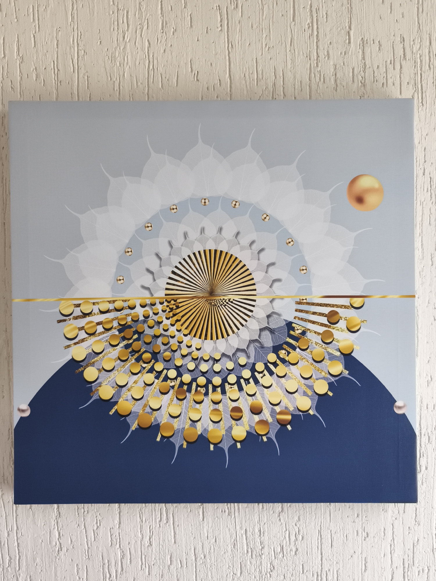 The image shows a square canvas hanging on a textured white wall. The canvas features an abstract design. The upper half of the canvas has a light blue-grey background with a large, translucent white circular design resembling stylized leaves. Embedded within this design are small gold-colored spheres. A gold-colored line horizontally bisects the canvas. Below the line, a dark blue semi-circular shape forms the background. Upon this dark blue field, numerous gold-colored circles are arranged, each connected to a translucent white, leaf-like extension, creating a radiating pattern. A larger, solid gold sphere is positioned in the upper right corner. Two pearl-like spheres are at the lower left and right edges of the dark blue semi-circle. At the horizontal center of the canvas is a radial design of parallel golden lines. No people or specific actions are depicted. The environment is a simple interior setting with a focus on the artwork. There is no indication that this image originates from or depicts a location in El Carmen, Mexico.