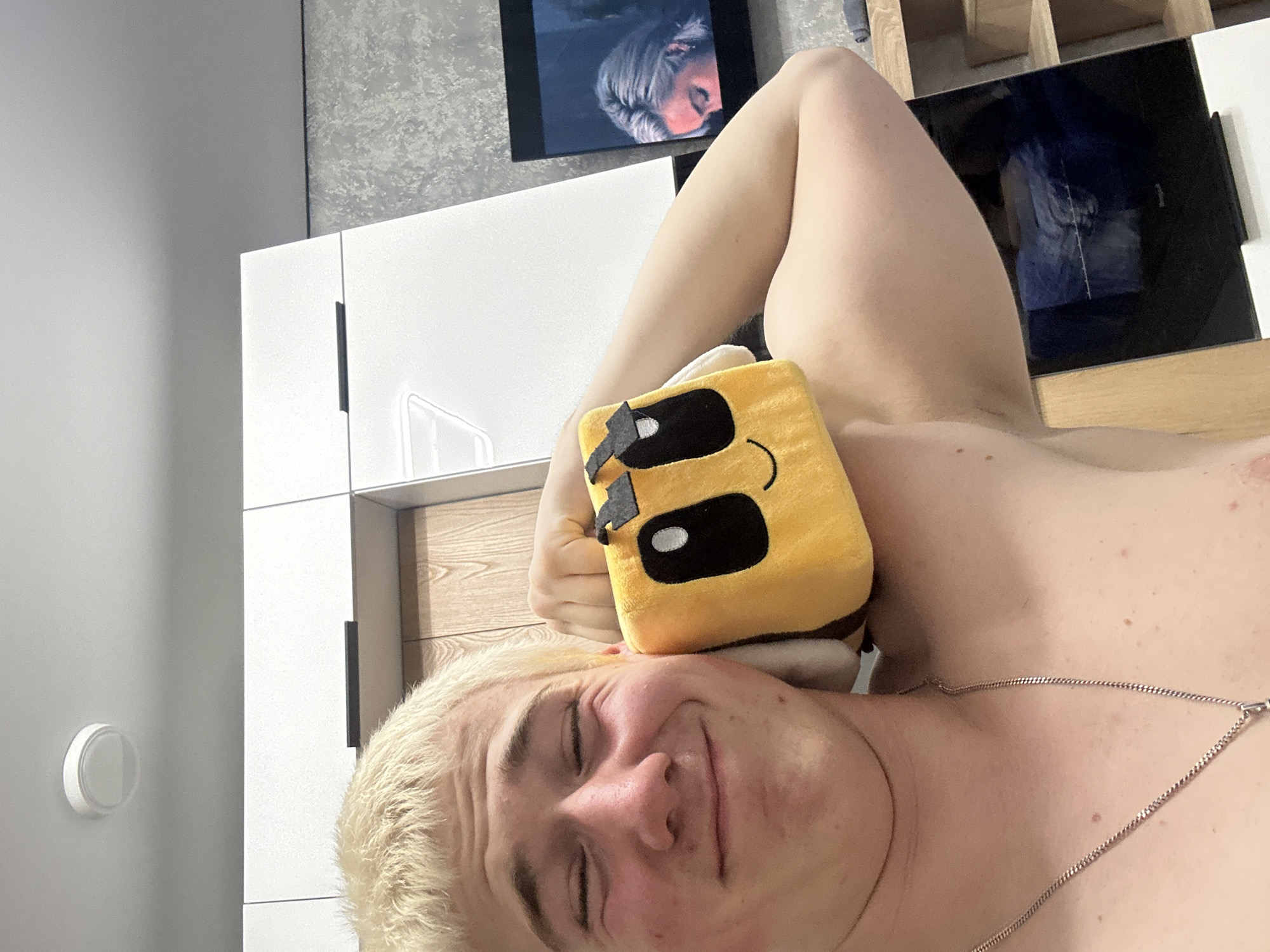 The image features a young man with short blonde hair, lying on his side and looking towards the camera with his eyes closed or squinting and a gentle smile. He appears to be shirtless, wearing a silver chain necklace. A yellow, square-shaped plush toy with large black eyes and a black smiling mouth (resembling a bee or a character from Minecraft) rests on his right arm, close to his head.
The setting is indoors, likely a modern bedroom or living area. In the background, there are white, sleek cabinets or furniture. Above these, a wall-mounted screen or tablet displays an image of a person with light-colored hair, possibly a reflection or a paused video. Further back, a wooden shelving unit is visible. On the far left, a white circular object, possibly a thermostat or light switch, is mounted on a plain grey wall. The overall ambiance is relaxed and comfortable. No visible text is present.