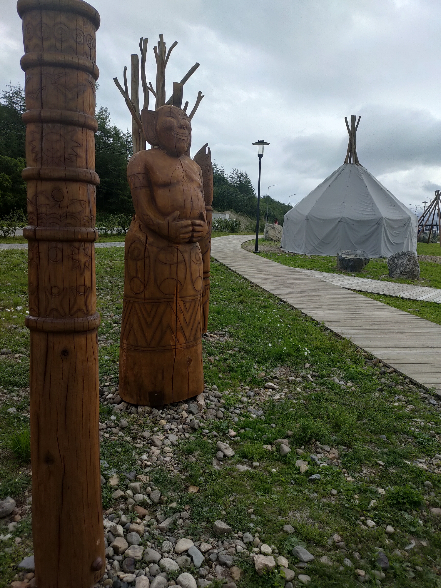 A wooden pole with carvings stands in the foreground to the left, its surface textured with rings and various symbols. To its right, a large wooden sculpture of a figure with a broad face and hands clasped at its waist is visible, also adorned with carved patterns. Behind these figures, a pathway made of wooden planks curves through a grassy area dotted with stones. In the mid-ground, a large, light-colored tent, possibly a yurt or tipi, is set up. Further in the background, trees cover a hillside under a cloudy sky. The scene is located in Magadan, Russia.