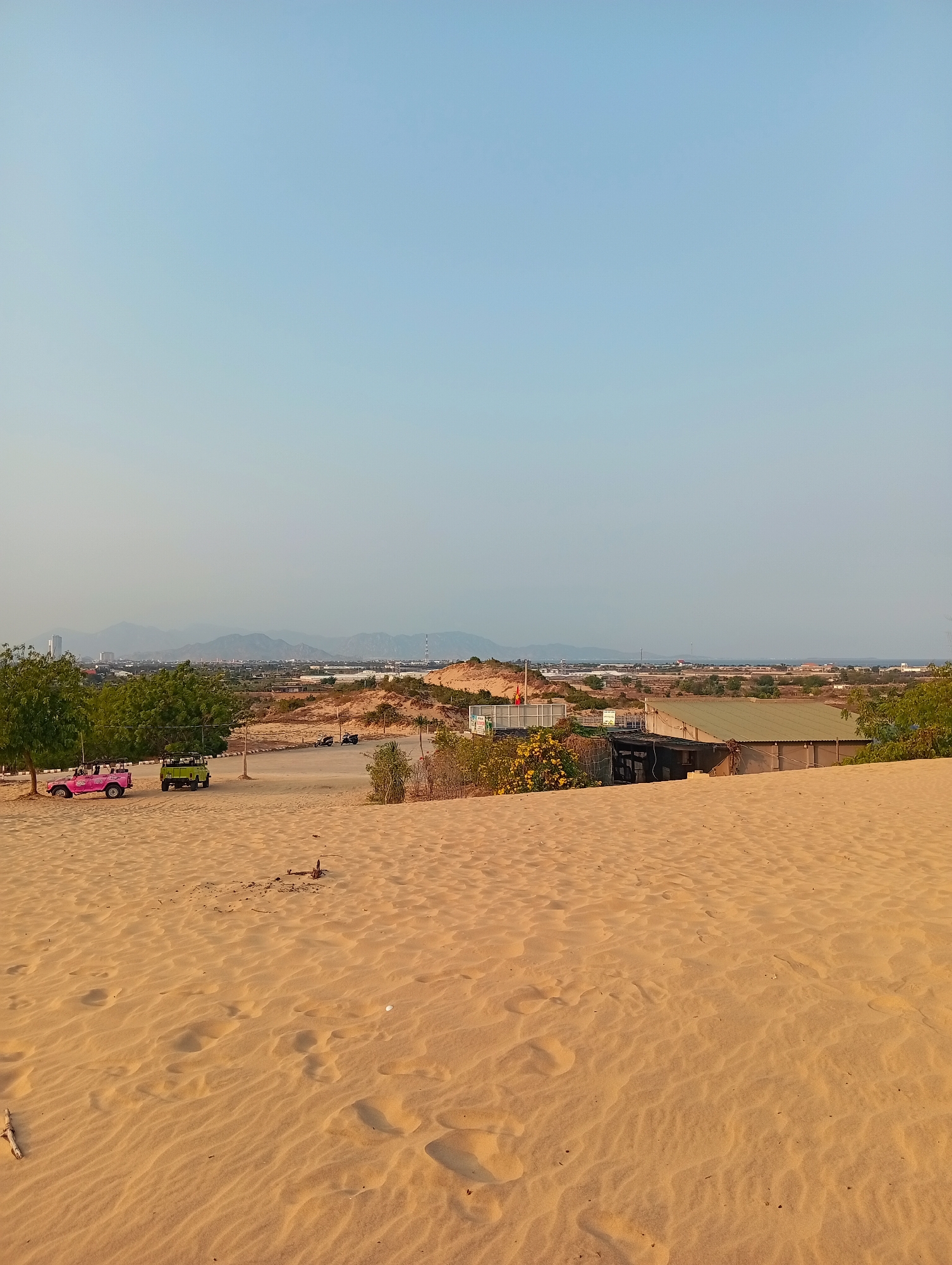 The image depicts a sandy landscape, possibly a sand dune, in Phuoc Dinh, Vietnam. The foreground consists of sand with wind-blown patterns. In the mid-ground, there is some vegetation and a building with a corrugated metal roof. Two open-top vehicles, one pink and one green, are parked under trees. In the background, a cityscape is visible with mountains in the distance under a clear sky.