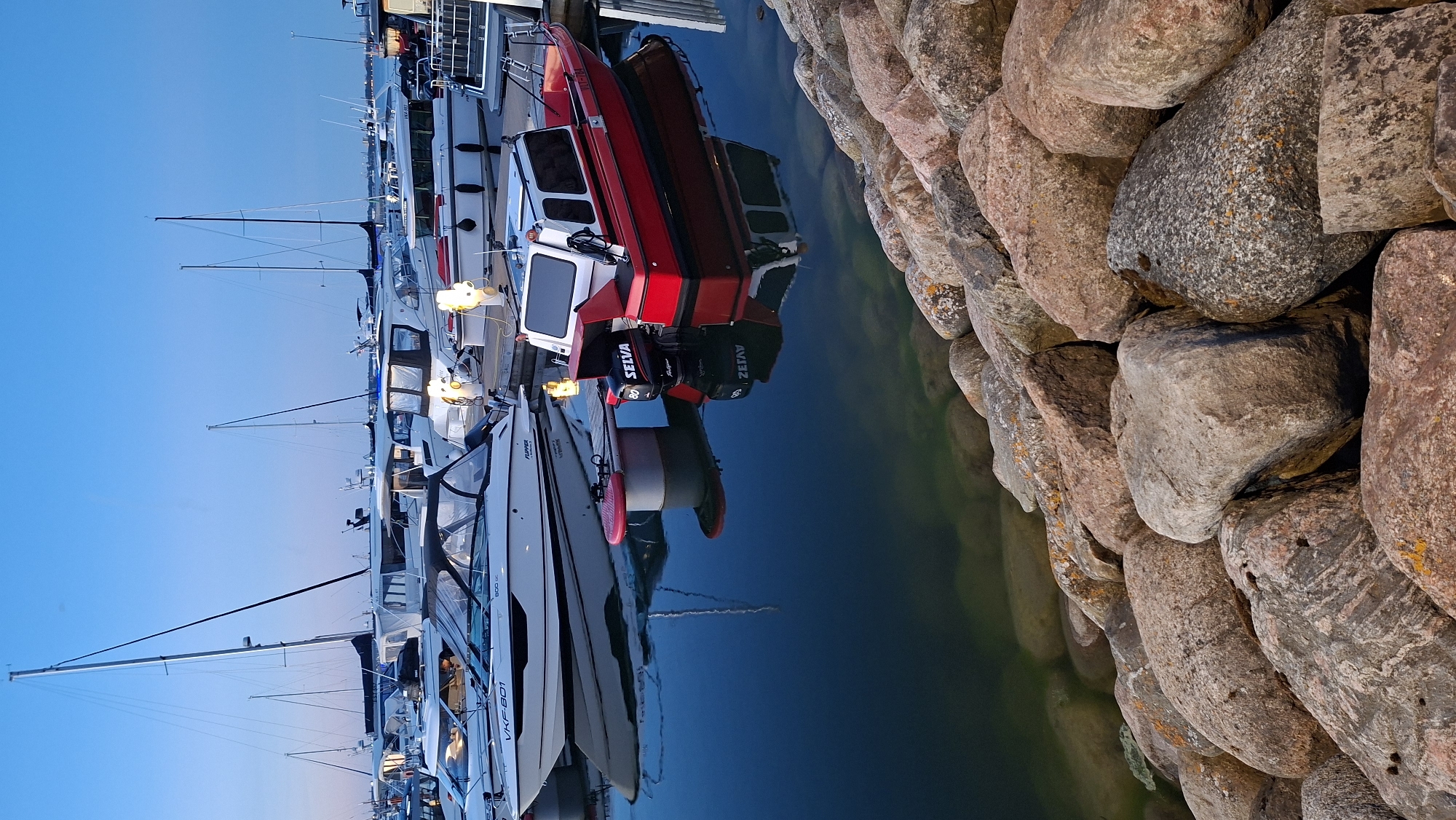 The image depicts a harbor in Tallinn, Estonia, likely at dusk. Several boats are docked, with a prominent red and white motorboat in the foreground reflecting in the dark water. Behind it, a white motorboat with a dark canopy is also visible. In the background, sailboats and other vessels are moored. The scene is framed by a rocky shoreline on the right side of the image. The sky is a clear, deep blue, suggesting the transition from day to night. No people are visible in the image.