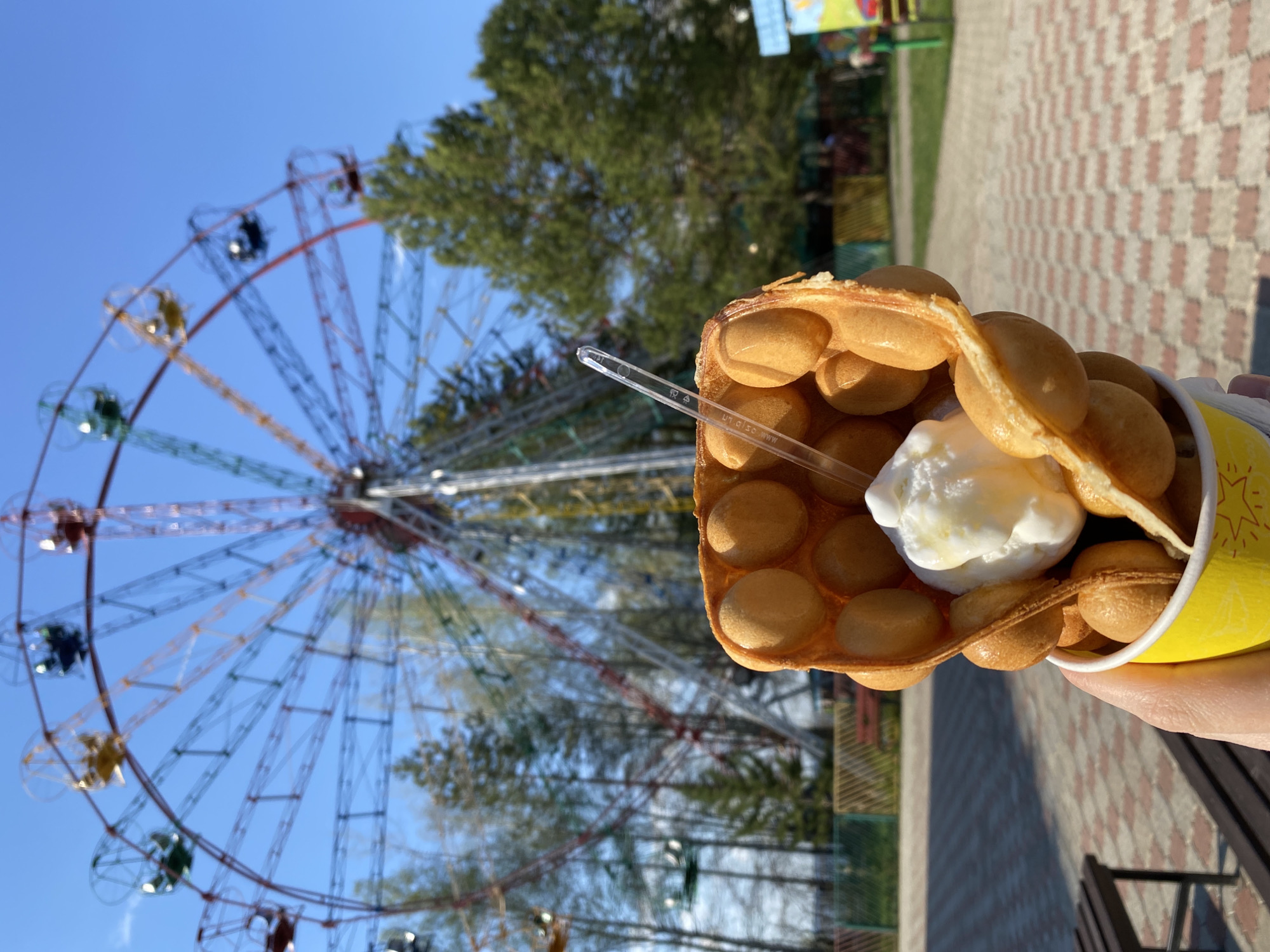 A person holds a bubble waffle with ice cream in front of a Ferris wheel. The waffle is in a yellow cup and has a clear plastic spoon sticking out. The Ferris wheel is large and has cars in various colors. The background includes trees and a paved area with patterned tiles. The location is likely Chaikovsky, Russia.
