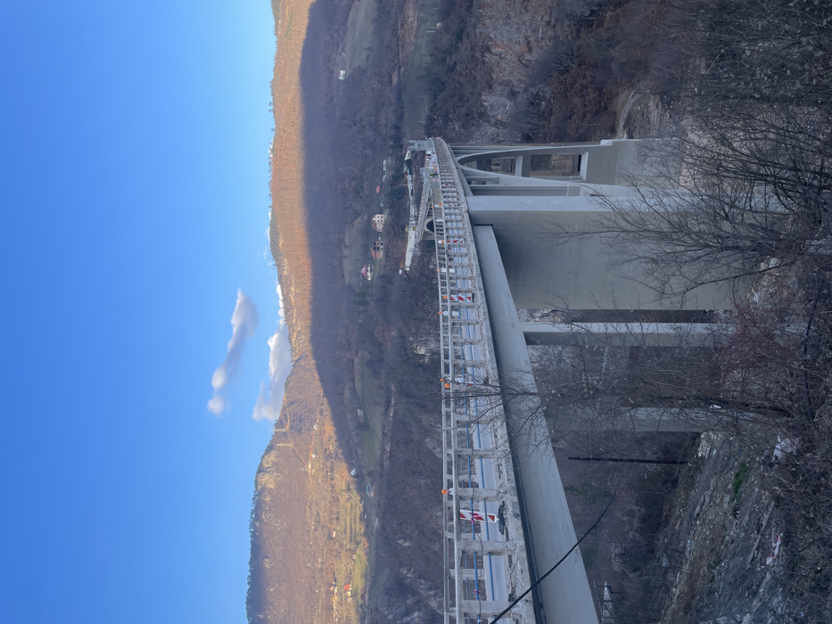 This image depicts the Đurđevića Tara Bridge, located in Đurđevića Tara, Montenegro. The scene is an elevated, panoramic view of the multi-arched concrete bridge spanning a deep, forested canyon under a clear, bright blue sky with scattered white clouds.
The bridge, constructed of light grey concrete, features a roadway marked by numerous orange and white striped traffic cones or bollards, indicating ongoing maintenance or construction. Several individuals, identifiable as workers by their orange high-visibility clothing, are present on the bridge deck. Blue scaffolding or netting is visible along the inner edge of the bridge's railing. The bridge's substantial concrete piers and arches descend into the valley below.
The surrounding environment consists of steep, mountainous terrain, predominantly covered with bare deciduous trees, suggesting a late autumn or winter season. The left mountain slope is brightly illuminated by sunlight, revealing scattered houses and utility poles. In contrast, the right mountain slope is largely enveloped in shadow. The immediate foreground features a sparse arrangement of bare tree branches. The bottom of the canyon is not directly visible from this perspective.