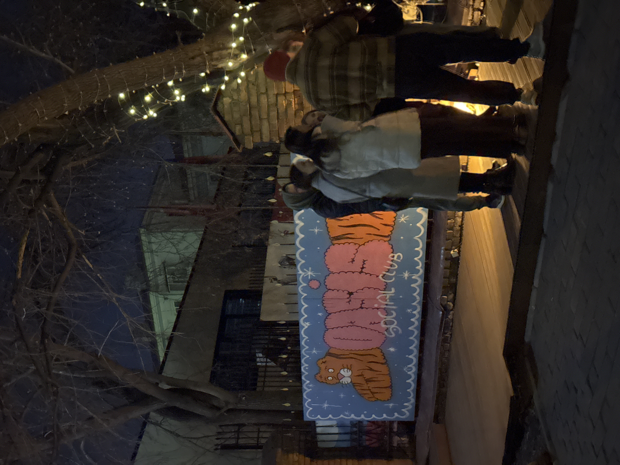 The image depicts a group of people standing near a sign that reads "Social Club" vertically, with cartoonish pink blobs above a tiger cartoon image, possibly the name of an establishment. The lighting suggests it is evening. A tree with bare branches and string lights is visible in the background. Based on research of the location of the "Social Club" signage this image is likely taken in Krasnoyarsk, Russia. The environment appears to be an urban sidewalk. People are conversing or looking at the sign.