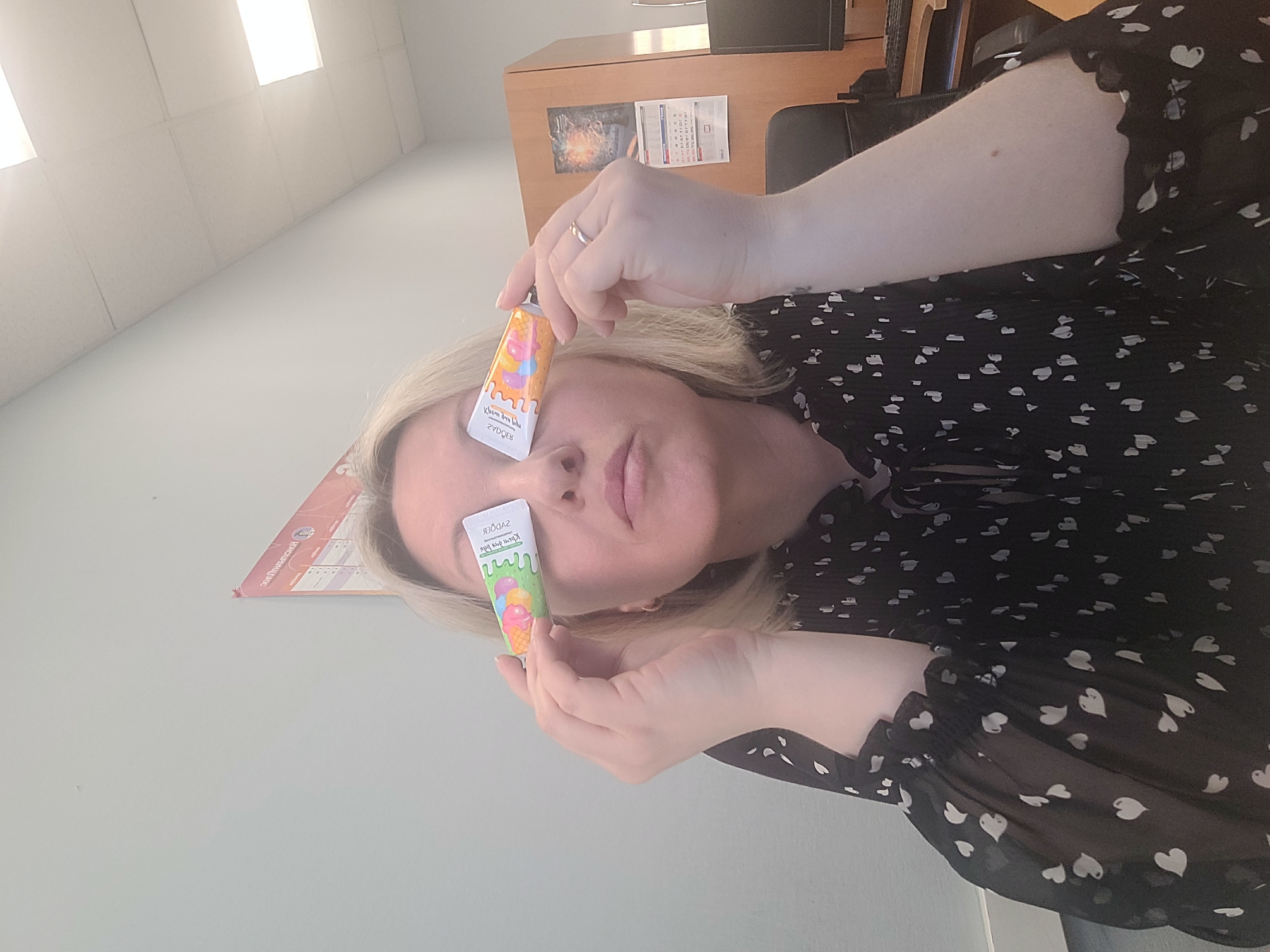 A woman with blond hair is holding two small tubes of children's hand cream in front of her face, one in front of each eye. The tubes are decorated with colorful images of ice cream cones. The woman is wearing a black blouse with white heart-shaped patterns. She is indoors, likely in an office setting, with a desk and computer monitor visible in the background. A calendar and a framed image are on the desk. Based on the text on the tubes written in Russian, the location is likely Izhevsk, Russia.