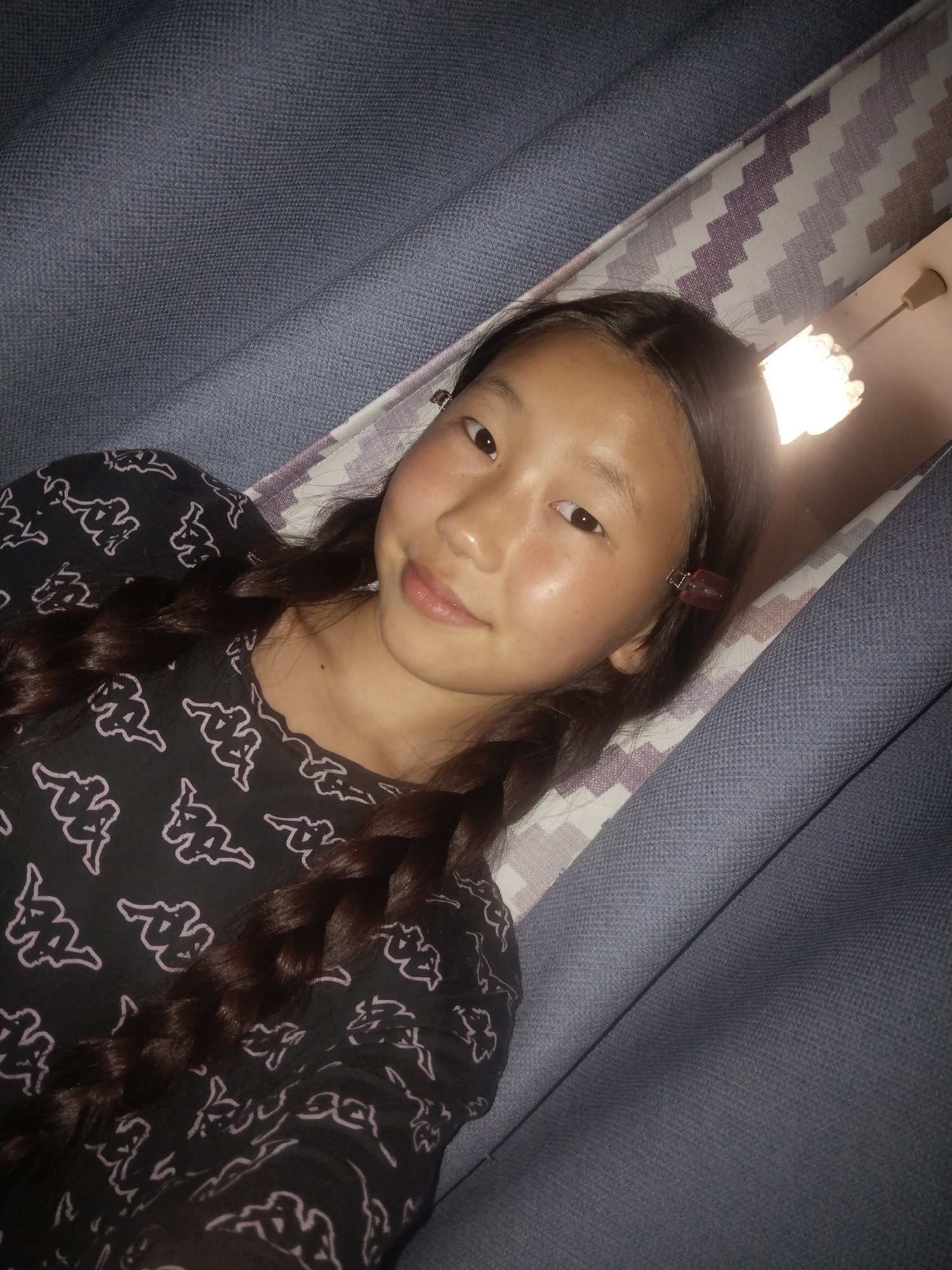 A young East Asian girl with brown hair in two braids is lying down. She is wearing a black shirt with a repeating white Kappa logo pattern. Her head is tilted slightly to the left, and she is looking towards the camera with a faint smile. A red hair clip is visible on the right side of her head. The background consists of textured purple and white fabric, along with some dark blue fabric. A light fixture is visible in the upper right corner, suggesting the image was taken indoors. There is no identifiable information that would place this image in the city of Ulan-Ude, Russia.