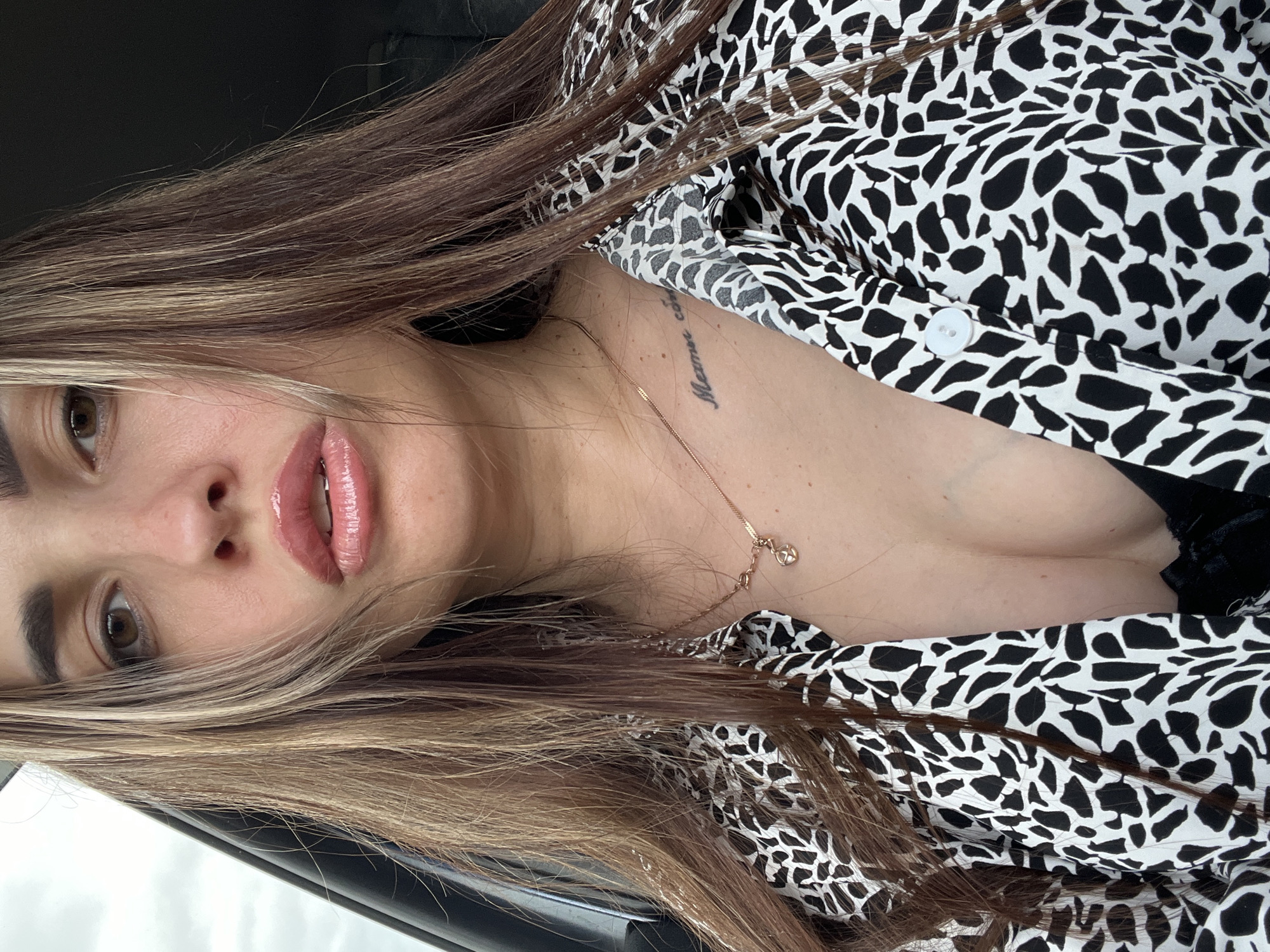 An adult female subject is centered in a close-up, chest-up shot. She has long, medium brown hair with blonde highlights, styled to fall over her shoulders and frame her face. Her gaze is directed towards the viewer. Her lips are full, covered in a clear gloss, and slightly parted. She is wearing a black and white abstract patterned button-down shirt, unbuttoned to reveal a black undergarment and cleavage. Two delicate gold-toned chain necklaces are visible: one with a small circular pendant on the left side of her chest, and another simpler chain further to the right. A script tattoo is located on her right clavicle, partially revealing Cyrillic characters, specifically "Мамин". The environment appears to be the interior of a vehicle, with a dark door frame visible on the far left and an overcast sky seen through a window beyond it. The background on the right is dark and indistinct, consistent with a car interior. The lighting appears natural, coming from the left. Location: Unknown, Unknown.