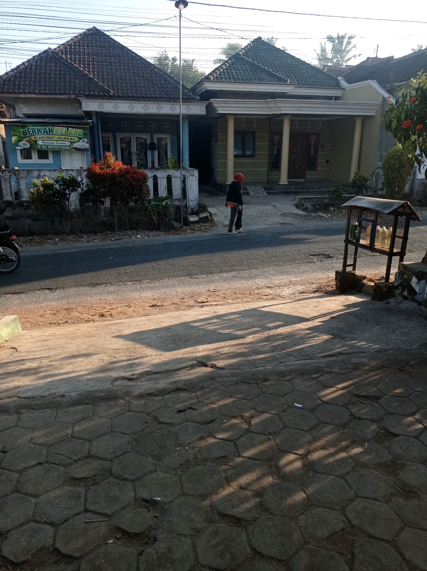 In Purwosekar, Indonesia, a street scene unfolds under a clear sky with the warm glow of the sun casting long shadows. Two houses stand side-by-side; the one on the left is painted light blue and features a sign advertising "BERKAH KELAPA" which offers grated coconut and coconut meat and takes orders via WhatsApp at 085103152062. Red and green foliage adds vibrant color to the front of this house. The house to the right is painted a pale green and has a more traditional Indonesian architectural style with prominent columns supporting the roof. A person wearing a red hooded jacket and dark pants stands in the middle of the asphalt road, facing away from the viewer and towards the green house. To the right of the road, a wooden stand with a wire cage houses several bottles, some appearing to contain liquids of varying colors, suggesting a small roadside stall. The foreground is paved with hexagonal concrete blocks, some of which are cracked and uneven. The street itself is asphalt, with a distinct dividing line of lighter concrete or gravel. A motorcycle is partially visible on the far left edge of the image. The overall impression is a quiet, everyday scene in a residential area.