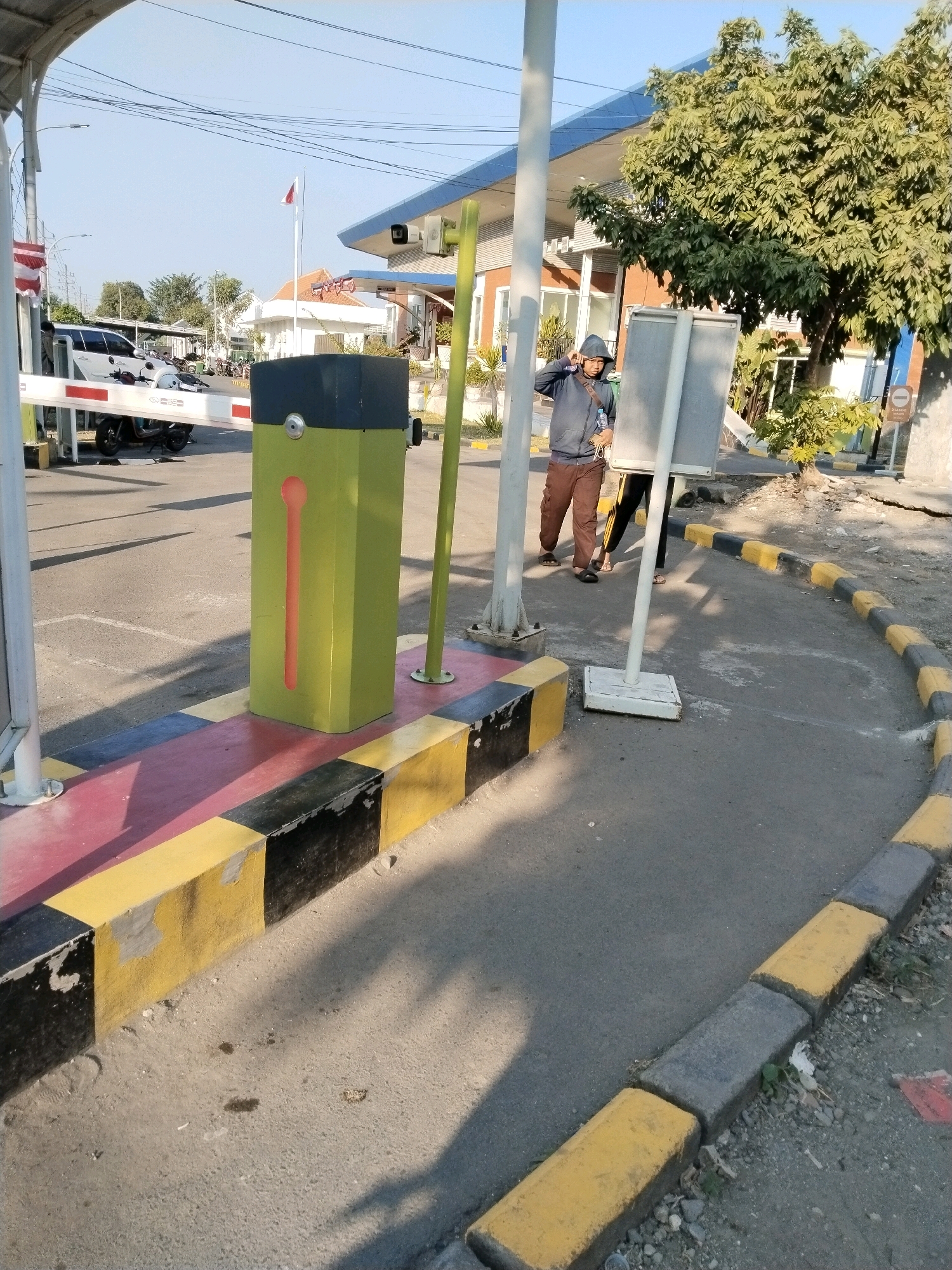 The image depicts an outdoor scene at what appears to be a checkpoint or entrance, possibly in Wonocolo, Indonesia. A white barrier gate is partially lowered, obstructing a paved road. To the left of the gate, a white car is parked. In the foreground, a bright green rectangular bollard with an orange vertical slot is positioned. The ground around the bollard is marked with alternating bands of red, black, and yellow paint.
Two individuals are visible on the right side of the image. One person, wearing a grey hooded jacket and brown pants, is standing near a gray sign on a white pole, appearing to be looking at their phone or holding it up. Another person, wearing black pants with yellow stripes and a grey top, is standing slightly behind and to the right of the first person. They are both facing towards the viewer.
The background shows a building with a blue awning and orange-brick-colored walls, possibly a commercial establishment or office. Trees with green foliage are present behind the building and to the right. The sky is clear and blue, suggesting a daytime setting. The overall environment appears to be a functional area with clear markings for traffic control and pedestrian pathways.