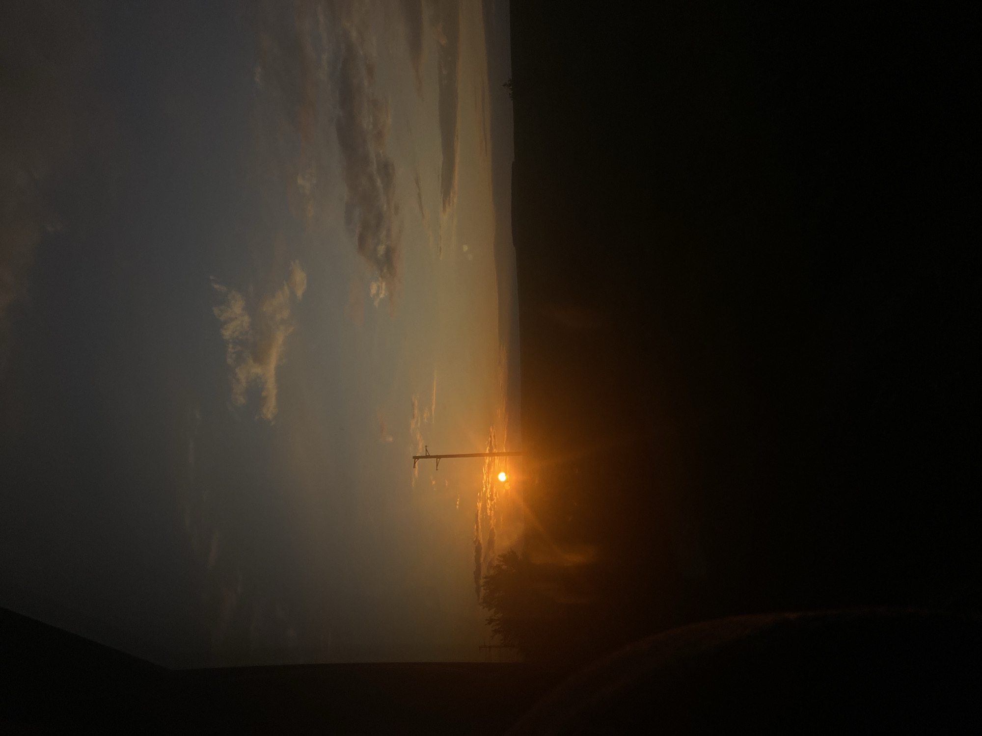 The image displays a dramatic sunset or sunrise over a landscape, likely in Buhovtsi, Bulgaria. The sun, a small but intensely bright and glowing orb, is positioned low in the sky, dominating the center-right of the frame with its powerful golden light. Its rays spread across the sky, creating a radiant halo effect and illuminating the edges of clouds.
A utility pole or similar tall structure stands prominently in silhouette directly in front of the sun, its dark outline contrasting sharply with the brilliant light. To the right of the pole and beneath the sun, silhouettes of trees or dense foliage are discernible along the horizon line.
The sky exhibits a gradient of warm colors, ranging from fiery oranges and yellows near the sun to softer grays and blues further away. Scattered clouds, some catching the warm light and others appearing as darker masses, add texture to the sky. A distant, dark horizon line, possibly indicating hills or mountains, stretches across the lower portion of the illuminated sky. The immediate foreground and a significant portion of the image's left side are plunged into deep shadow, appearing almost black, with minimal discernible details.
The scene captures the serene and stark beauty of either dawn or dusk, characterized by strong contrasts between light and shadow. There is no visible text in the image.