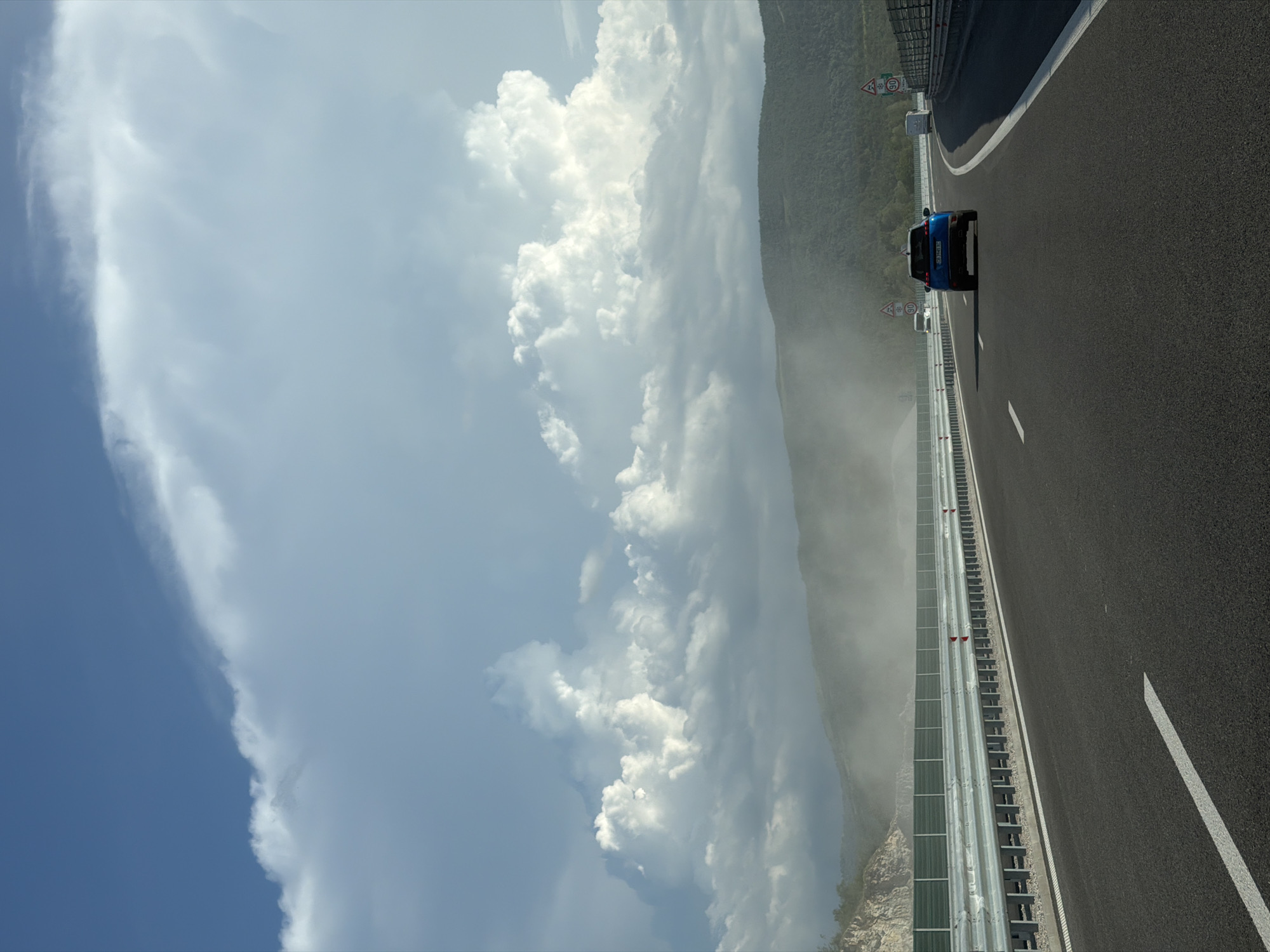 The image captures a perspective from a vehicle traveling on a modern highway. The road surface is asphalt with clear white lane markings. On the right side of the highway, a metal guardrail with a green sound barrier runs along the edge, with traffic signs visible in the distance. On the left side, another guardrail and sound barrier are present, bordering a rocky, excavated slope. In the background, a range of green, forested hills are shrouded in a hazy mist or dust. The sky is a mix of deep blue and dramatic, white cumulus clouds, with some darker, more ominous clouds towards the horizon. A dark blue SUV is visible in the distance, traveling in the same direction as the camera. Other vehicles, including a white van and a smaller white car, are also present on the highway.