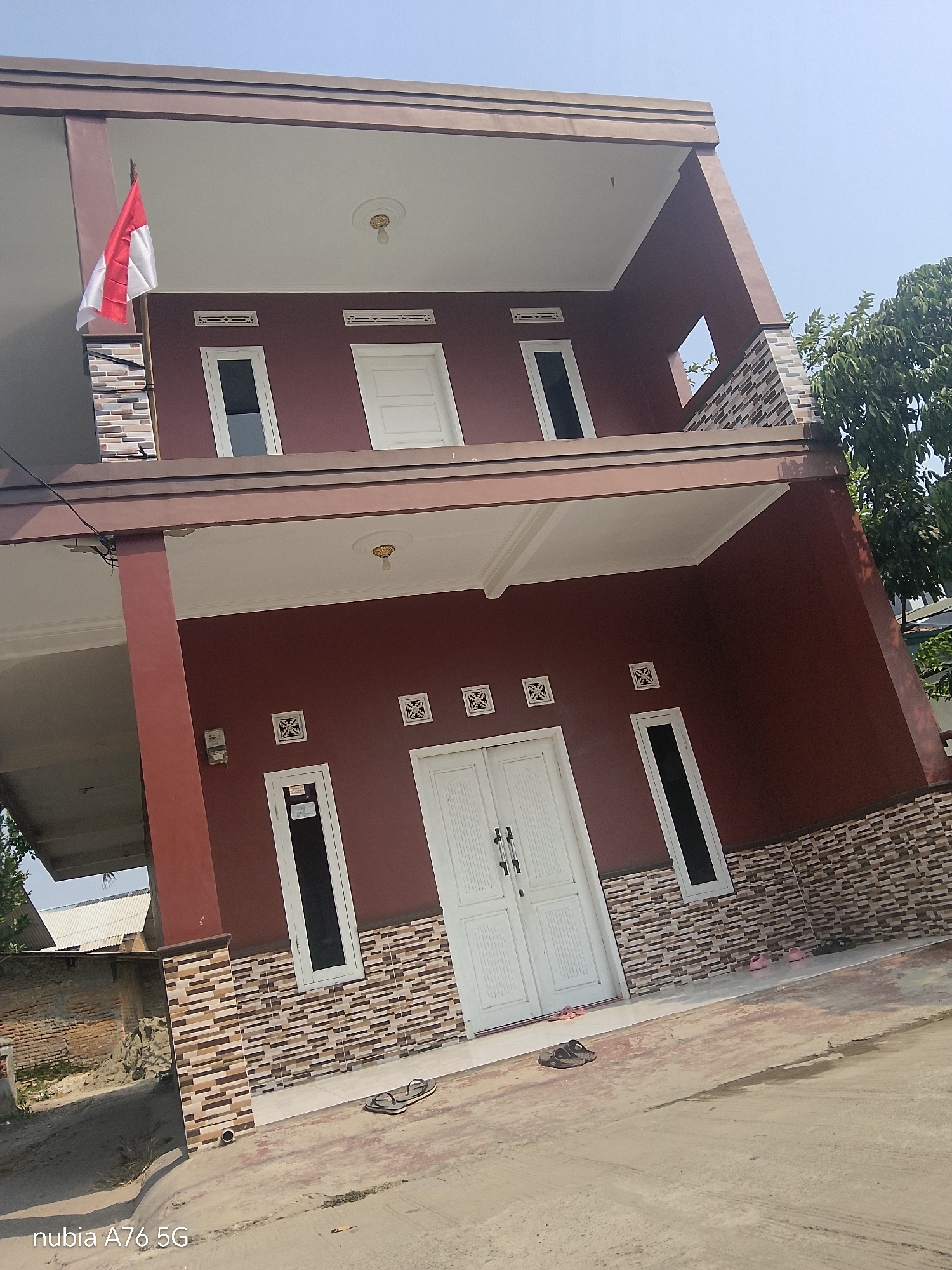 The image depicts a two-story house with a brown exterior, situated in Cakung, Indonesia. A red and white Indonesian flag is visible on the upper floor, to the left of the house. The ground floor features a double white door and several windows. The house is surrounded by a tiled facade and has a paved area in front. Two pairs of sandals are visible on the ground. The sky is clear and blue, with a tree visible on the right side of the image.
