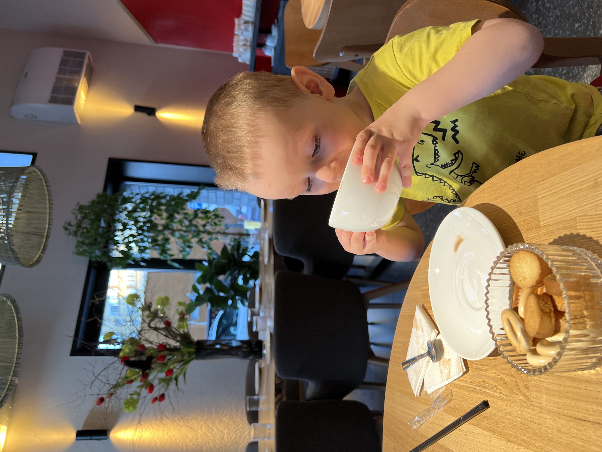 A young boy in a yellow t-shirt is drinking from a white cup at a wooden table in what appears to be a cafe or restaurant in Mytishchi, Russia. Next to him are a white saucer, a stack of napkins with a spoon, and a ribbed glass container filled with small bagels or biscuits. A black straw and plastic packaging are also on the table. In the background, there are other tables and chairs, a window with greenery, and a decorative floral arrangement. A white air conditioning unit is visible on the wall. The overall lighting suggests it is daytime.