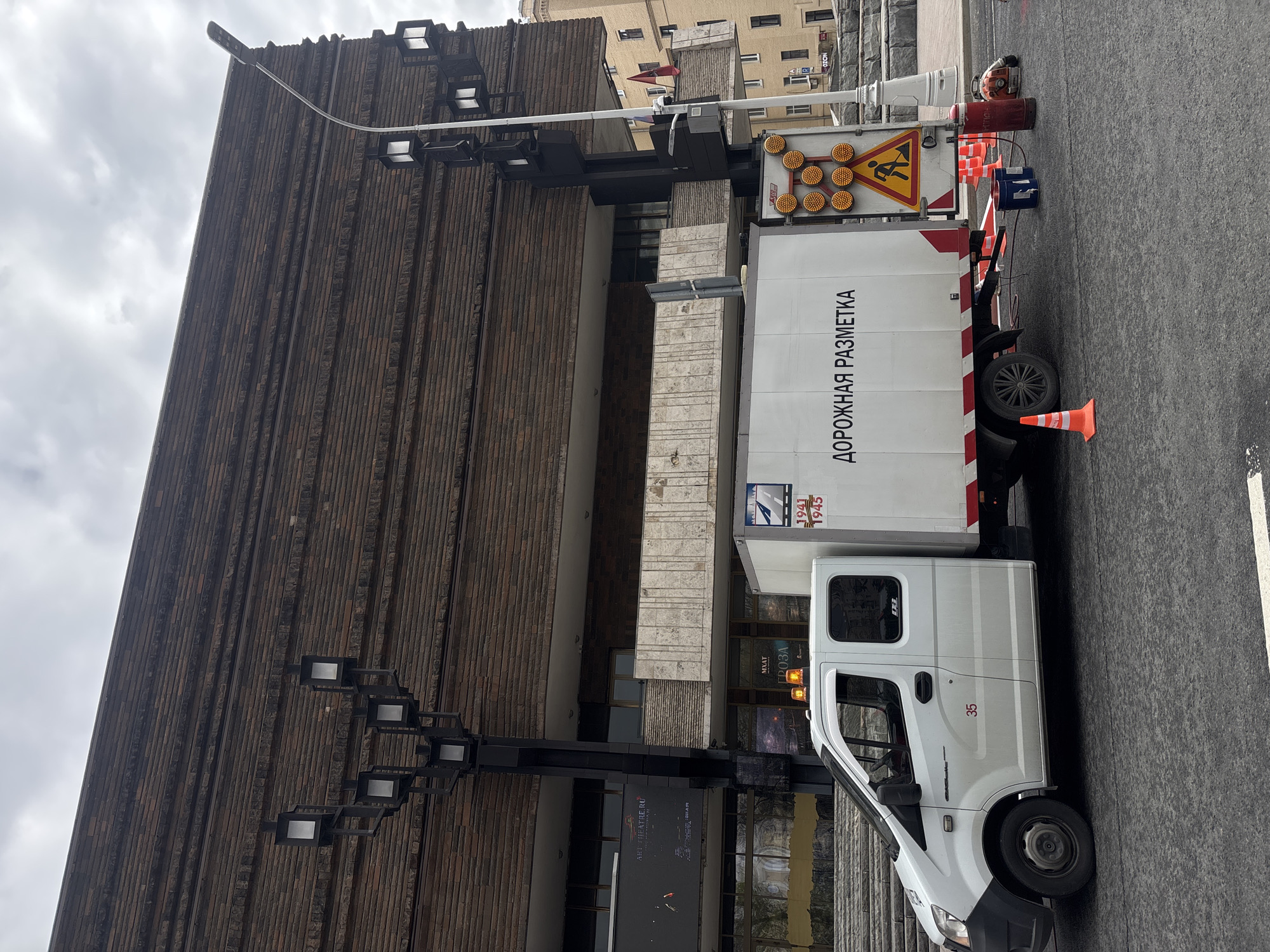 The image captures a street scene in Moscow, Russia, featuring a white utility truck parked adjacent to the distinctive façade of the Moscow Art Theatre (MXAT) named after Maxim Gorky. The truck, labeled "ROAD MARKING" in Russian, is equipped with traffic control devices, including orange cones and a flashing arrow board. A man can be seen near the rear of the truck, appearing to be engaged in road marking activities. The architecture of the theatre is characterized by its vertical ribbing and modernist style. The sky is overcast, suggesting a cloudy day.