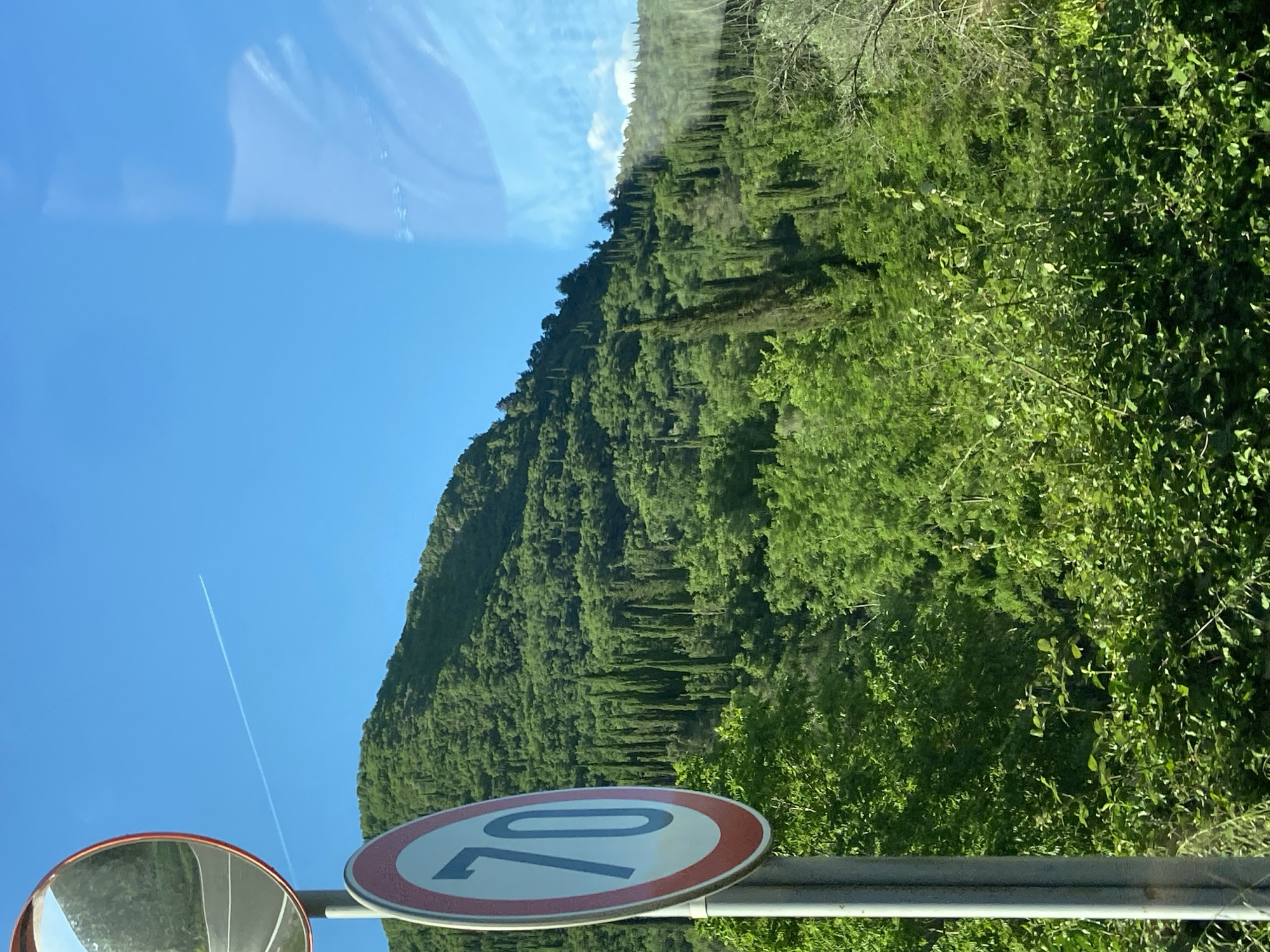The image depicts a scene in Sutorina, Montenegro. A lush, green hillside densely covered with trees dominates the background. In the foreground, a circular speed limit sign with the number "70" is visible. To the left of the sign is a convex mirror, also circular, reflecting the road. The sky is clear and blue. A vapor trail from an aircraft is visible across the blue sky.