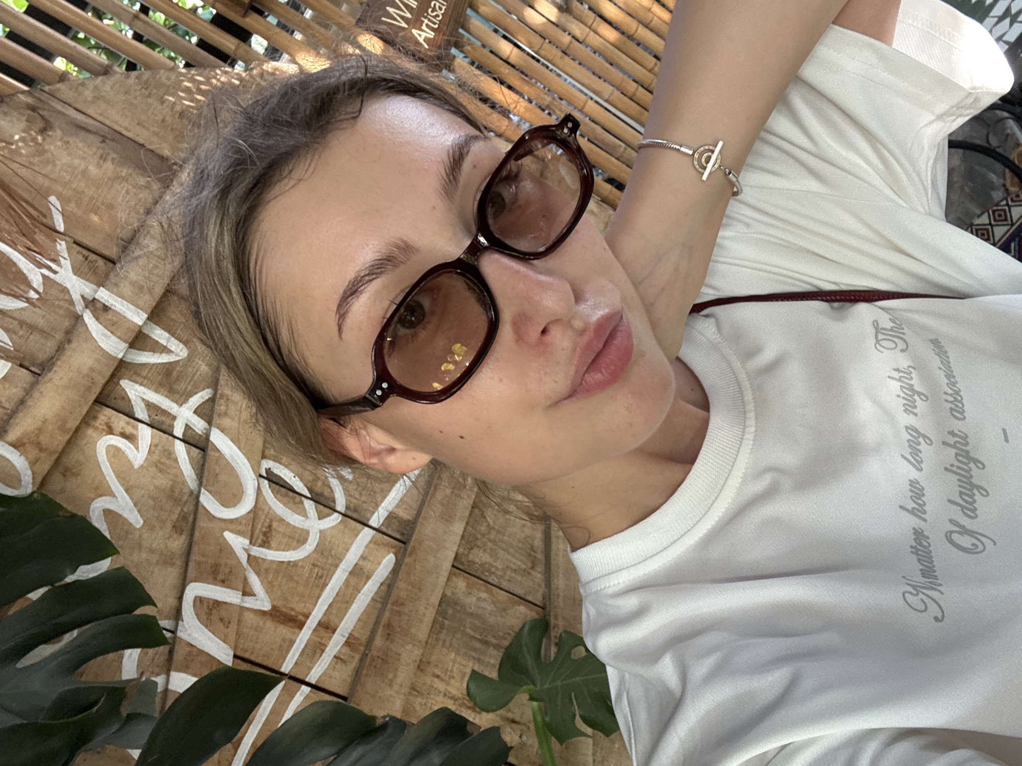 A young Caucasian woman wearing sunglasses and a white t-shirt is taking a selfie. She is leaning her head on her hand and her eyes are looking at the camera. The t-shirt has some text on it, but it is partially obscured by the woman's arm. The background is a wooden wall with some plants and bamboo. There is also some writing on the wooden wall that appears to be in white paint. There is a bracelet on the woman's wrist. The image appears to have been taken outdoors in a cafe or restaurant setting. The lighting suggests it is daytime.