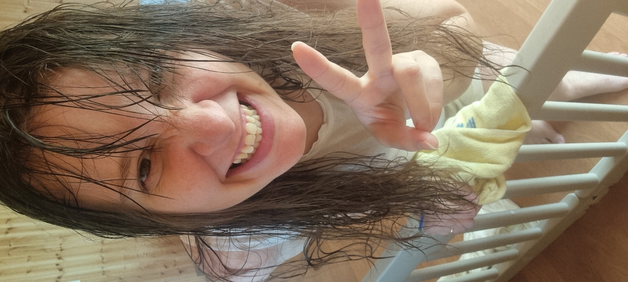A person with wet, dark hair is smiling and making a peace sign with their right hand. The person's face is angled towards the camera. They are wearing a light-colored garment. In the background, there is a wooden floor and a white, slatted object, possibly a piece of furniture or a crib. A yellow cloth with a blue logo is draped over the white, slatted object. The location of the image is Dunnae-myeon, South Korea.