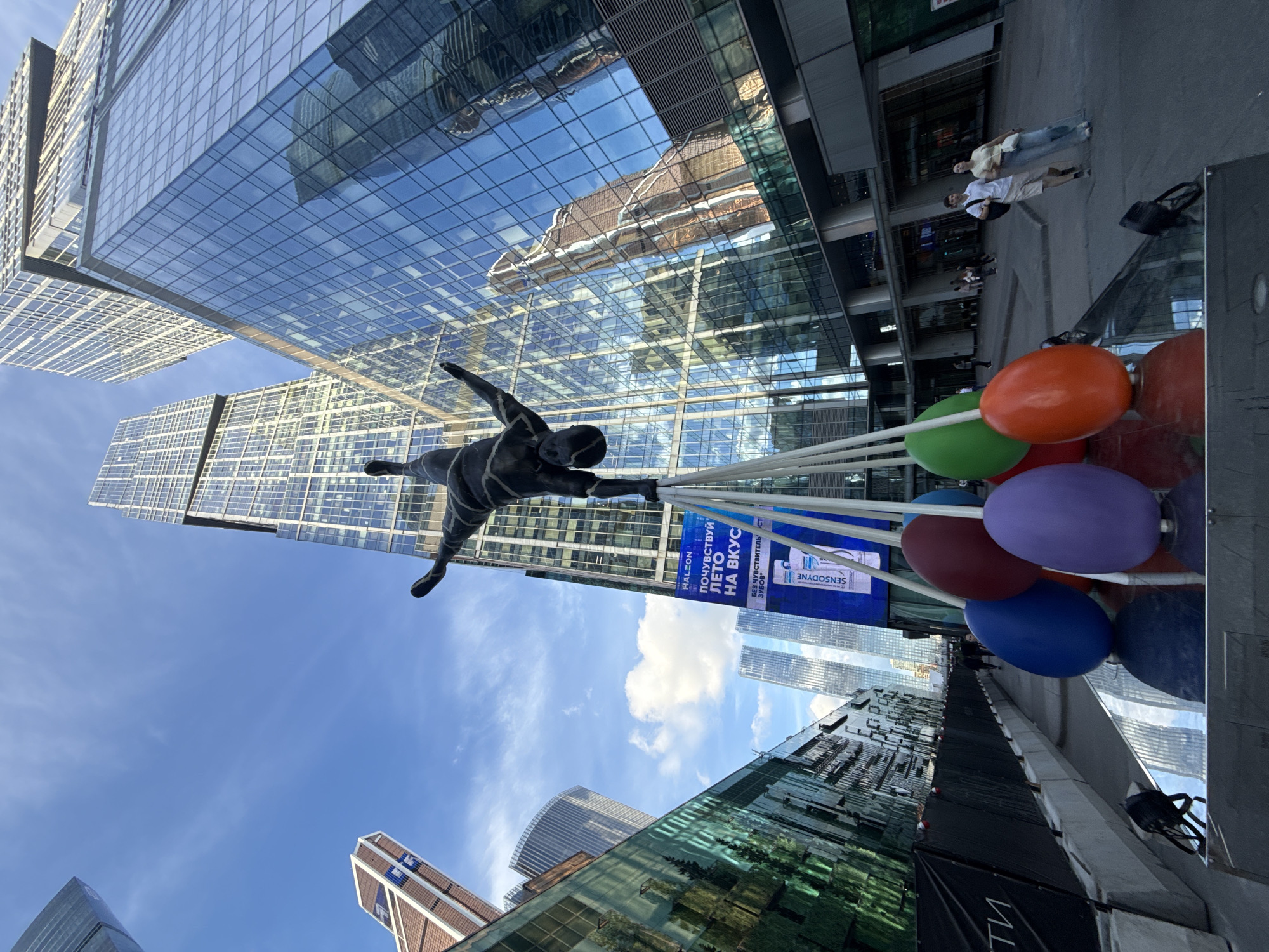 The image captures an outdoor urban scene with a unique art installation in the foreground. A black, human-like sculpture is depicted in a dynamic pose, appearing to fly or leap, supported by a cluster of colorful balloons. The balloons are red, orange, green, purple, and blue, and they are tethered to the sculpture by white strings.
The setting is a modern city with towering glass skyscrapers that reflect the sky and surrounding buildings. The perspective is looking upwards, emphasizing the scale of the architecture. In the background, several people are visible walking on a paved area, and on a ledge of one of the buildings, there is a billboard.
The billboard displays text in Cyrillic script. One prominent line reads "ПОЧУВСТВУЙ ЛЕТО НА ВКУС" which translates to "Taste Summer". Below that, another line visible is "БЕЗ ЧУВСТВИТЕЛЬНОЙ". The brand "SENSODYNE" is also clearly visible on the billboard.
The sky is a clear blue with a few scattered white clouds, suggesting daytime, likely a bright and possibly warm day. The overall atmosphere is contemporary and vibrant, highlighting the interplay between art, architecture, and the urban environment. The angle of the shot and the reflections in the glass buildings create a sense of depth and visual interest. The presence of people adds a human element to the otherwise monumental structures.
