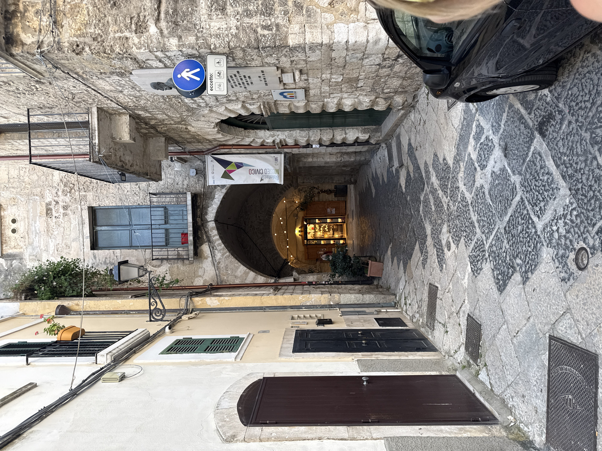 The image displays a narrow, historic cobbled alleyway flanked by multi-story stone and stucco buildings. The ground features a distinctive pattern of light and dark grey cobblestones. On the right, a portion of a black car is visible, parked alongside the street.
The architecture suggests an old European town, with varying window styles, some featuring green shutters and others metal grates. Utility cables are strung along the building facades. On the left, a decorative street lamp is attached to a wall, and a potted plant with red flowers sits on a window ledge.
Several signs are prominent:
- A circular blue sign with a white pedestrian symbol indicates a pedestrian zone, with text "eccetto:" (Italian for "except:") followed by small icons for bicycles and scooters, suggesting they are permitted.
- Below this, a white plaque with unreadable Italian text is mounted on the stone wall.
- A large white banner hangs across the alley's entrance, displaying "MUSEO CIVICO" (Civic Museum) and "Biblioteca" (Library) along with a colorful geometric logo.
- Further down the alley, an arched passage leads to an illuminated interior space, possibly a shop or part of the museum, with visible shelves and display items. String lights adorn the inside of the archway.
Manhole covers are embedded in the pavement, with one clearly bearing the Italian inscription "ACQUEDOTTO PUBBLICO" (Public Aqueduct). The scene appears tranquil, suggesting a daytime setting, possibly late afternoon, with no specific activity taking place. The visible Italian text, architecture, and pedestrian signage strongly indicate a location in Italy.