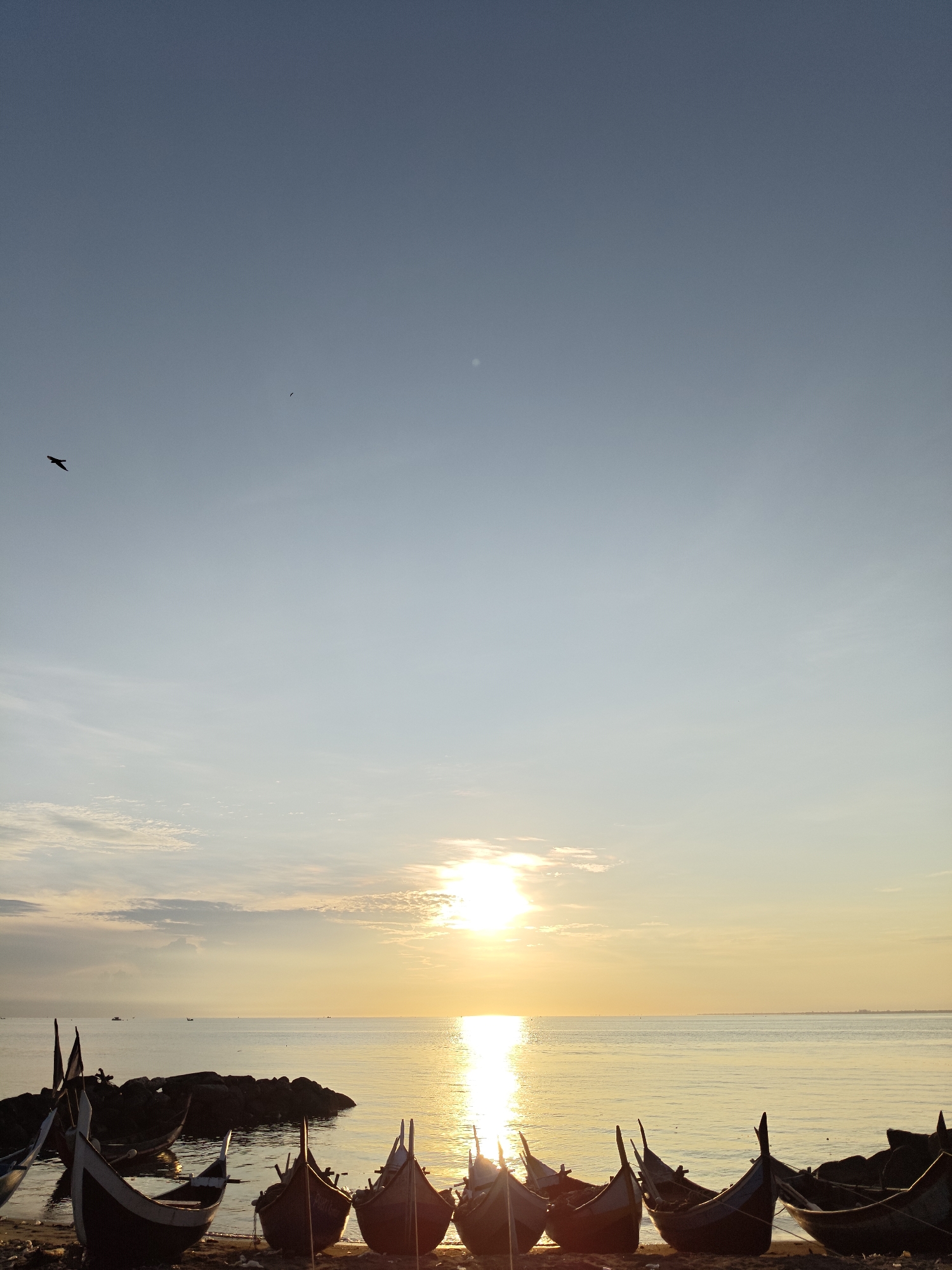 The image depicts a sunrise scene on a beach near Lhokseumawe, Indonesia. The sun is rising over the ocean, casting a bright reflection on the water. Several traditional fishing boats are lined up on the shore. A bird is flying in the upper left of the sky. The sky is clear with a few clouds near the horizon. The beach has a dark, sandy appearance. Rocks are partially submerged in the ocean near the boats.