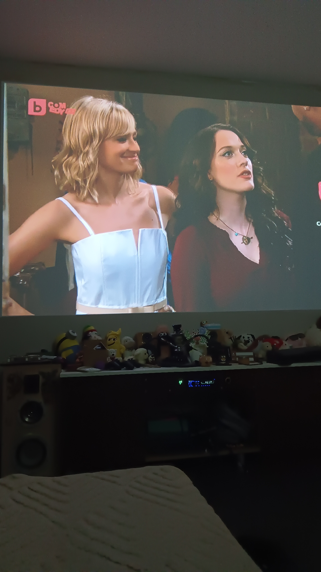 The image shows a television screen displaying a scene from a comedy TV show, viewed from an indoor setting. On screen, two women are prominent. The woman on the left has blonde, wavy hair with bangs, and is smiling. She wears a white camisole-style top with thin straps and a V-shaped cut-out in the front. The woman on the right has long, dark, wavy hair and is looking slightly upwards with a thoughtful or surprised expression. She wears a dark red V-neck top with a necklace featuring a heart pierced by an arrow pendant. In the top-left corner of the screen, a logo reads "b COMEDY HD".
Below the TV, an entertainment unit or shelf holds numerous plush toys and figurines, including a Minion, Mr. Potato Head, a yellow emoji character, small animal plushies, and a robot. To the left, part of a light-colored speaker is visible. An electronic device with green and blue lights is situated on the unit. The immediate foreground shows a textured, light-colored blanket or bedding, suggesting a cozy, relaxed atmosphere. The room appears dimly lit, with the TV screen being a primary light source.