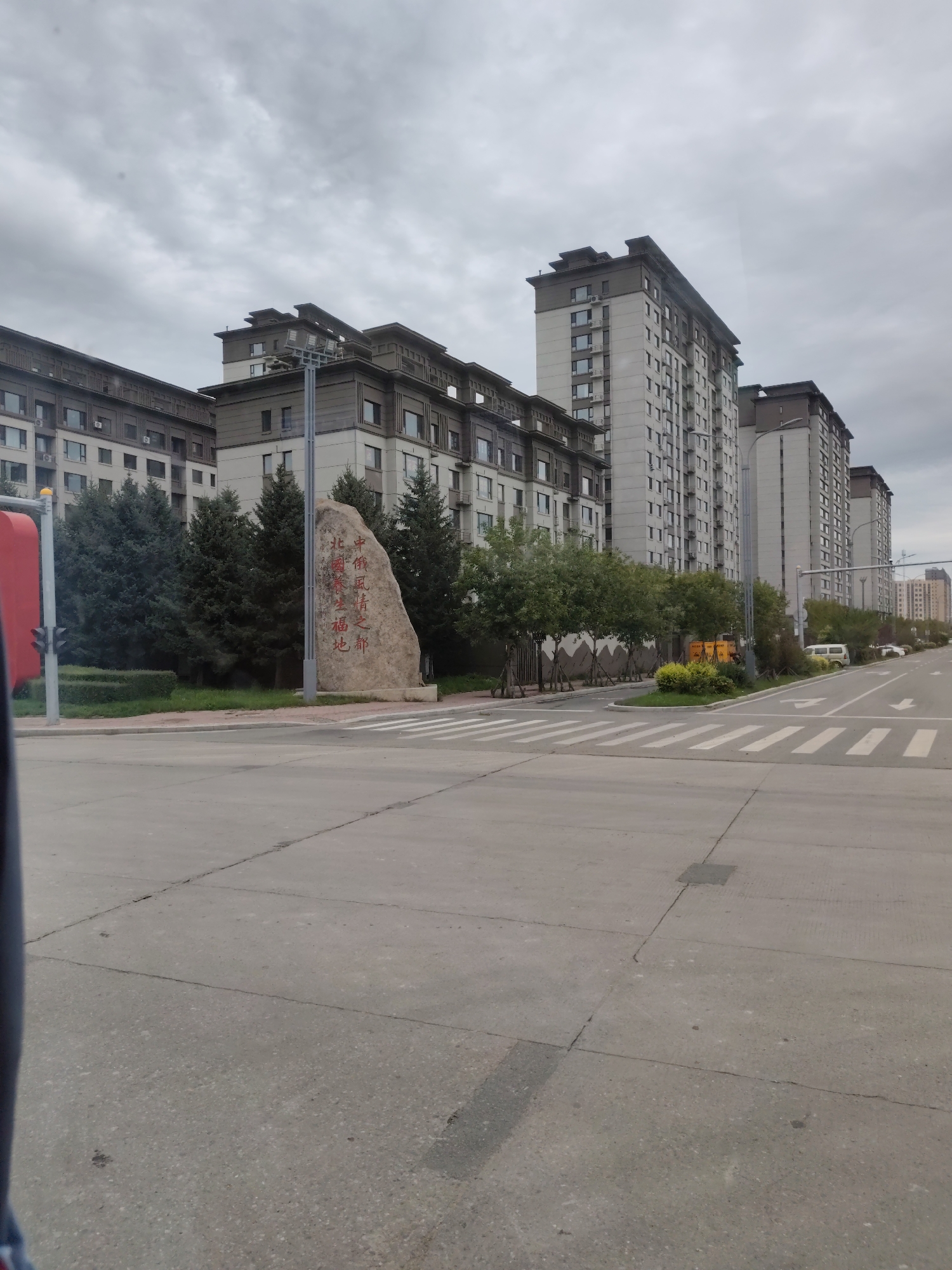 This image, taken in Heihe, China, shows a street scene with several modern, multi-story apartment buildings. In the foreground, a wide paved road with crosswalk markings dominates the scene. A large, rough-hewn rock with red Chinese characters inscribed on it stands to the left of the road, near some evergreen trees and manicured hedges. A traffic light pole with a red housing is visible on the far left edge of the image. The sky is overcast and gray. The architecture of the buildings suggests a residential development.