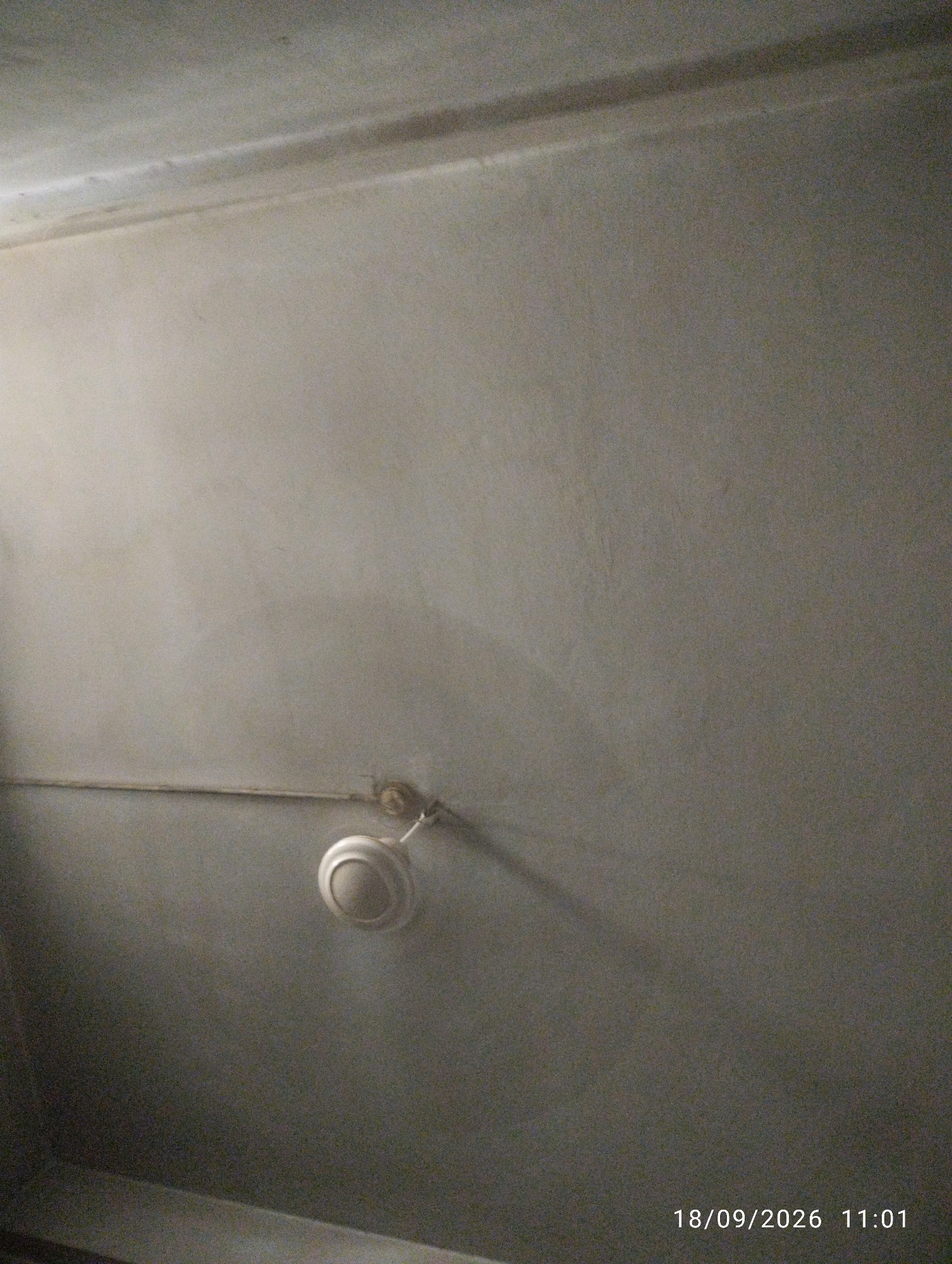 The image shows a ceiling with a circular fan attached. The ceiling is painted a light gray color and appears to be textured. There is a faint shadow cast on the ceiling from the fan, suggesting a light source from above and to the left. A thin, horizontal line runs across the ceiling, possibly indicating a seam or a conduit. The fan itself is white and has a central dome-shaped body with what appears to be a light fixture or a decorative element on top. A thin rod connects the fan to its mounting point on the ceiling. The timestamp "18/09/2026 11:01" is visible in the bottom right corner of the image, indicating the date and time the photograph was taken. The scene is indoors, likely in a residential or commercial space. There is no indication of any people or specific location within Chattogram, Bangladesh, other than the timestamp.