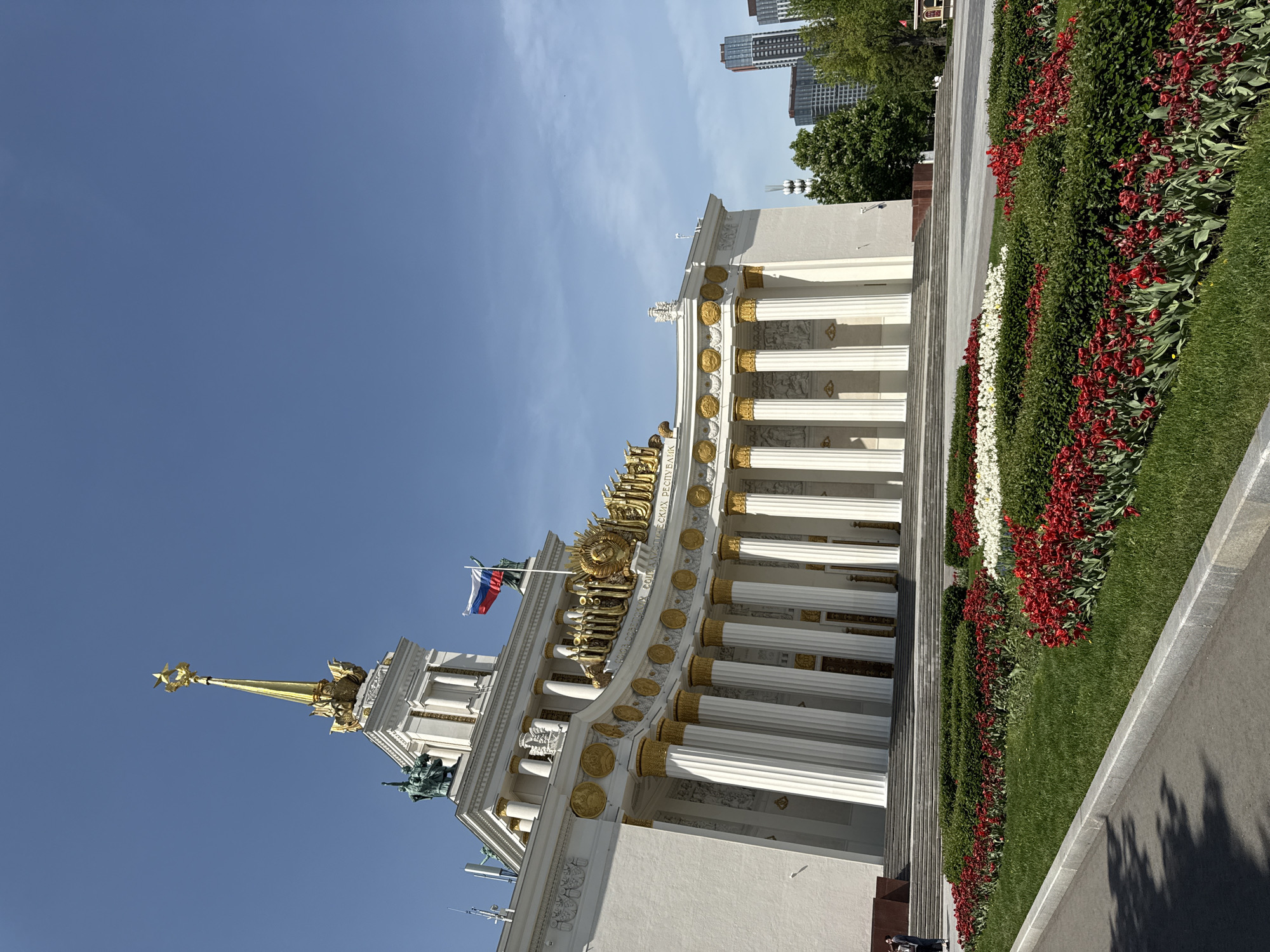 The image shows the Central Pavilion at VDNKh (Exhibition of Achievements of National Economy) in Moscow, Russia. The pavilion is a grand, ornate structure with white columns, gold decorative elements, and a spire topped with a star. A Russian flag flies atop the building. The foreground features a flower bed with red and white flowers, as well as a paved walkway. In the background, modern buildings rise above the trees. The sky is clear and blue.