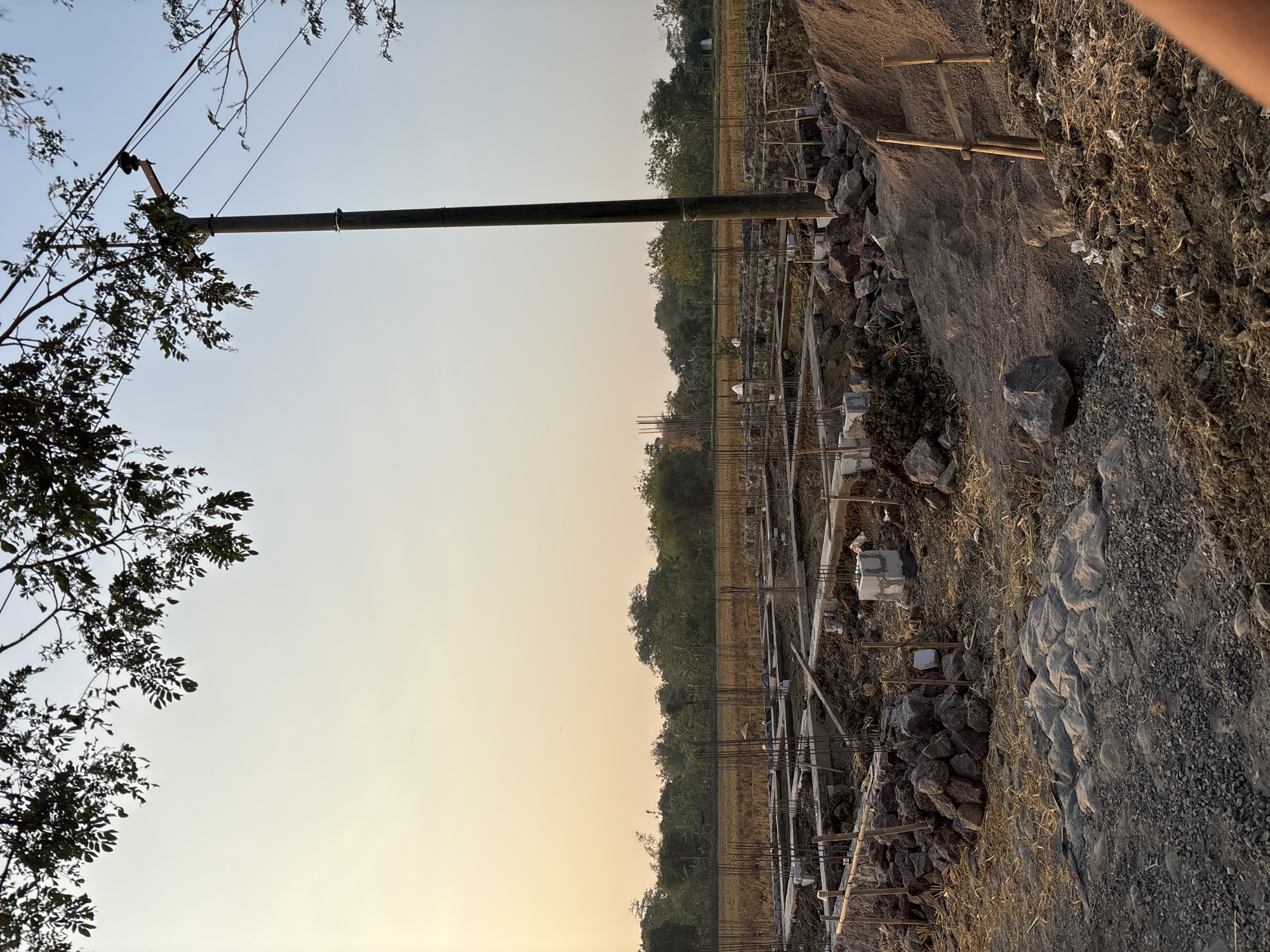 An outdoor, low-angle, medium-shot captures a construction site in Panongan, Indonesia, at dusk. The sky is a pale orange-yellow, transitioning to a lighter blue as it reaches higher. In the foreground, the foundation of a building is under construction, with concrete footings and rebar visible. Piles of rocks and sand are scattered around the site, along with some straw-like material and what appears to be a gray tarp. Wooden stakes and other construction materials are also present. A tall, dark utility pole stands to the right of the frame, with wires running from it. In the background, a field of dry vegetation stretches towards a line of trees. A few indistinct figures can be seen in the distance on the construction site. The overall impression is one of ongoing construction in a rural or semi-rural setting during the evening.