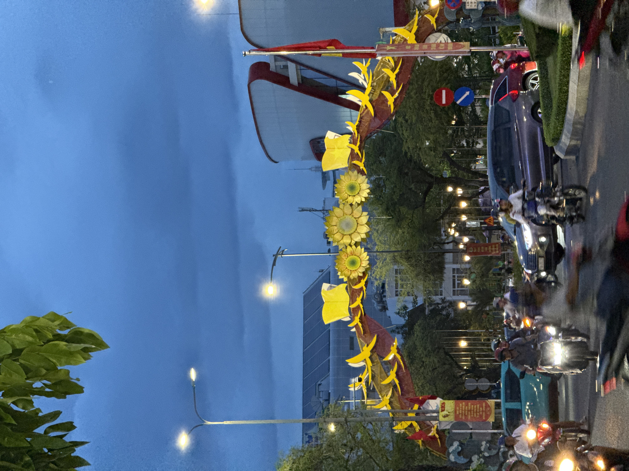 This image depicts a street scene at dusk in Nha Trang, Vietnam. A large, decorative archway adorned with yellow sunflowers, yellow bird-like figures, and red banners spans across the road. The archway is illuminated by streetlights, and the sky above is a deep blue, indicating the transition from day to night.
The street is busy with traffic, primarily motorbikes, with a few cars also visible. Several people are riding motorbikes, wearing helmets. Some vehicles appear to be in motion, creating motion blur in the image. Buildings can be seen in the background, including a modern structure with a curved red and white facade, and another building with a dark-tiled roof that has solar panels. Trees line the street, and streetlights are also present, some of which are lit. Traffic signs, including a red circle with a horizontal bar and a blue arrow pointing down, are visible on poles. Red flags are also displayed on a pole near the archway. The overall impression is of an urban environment decorated for a festive occasion.