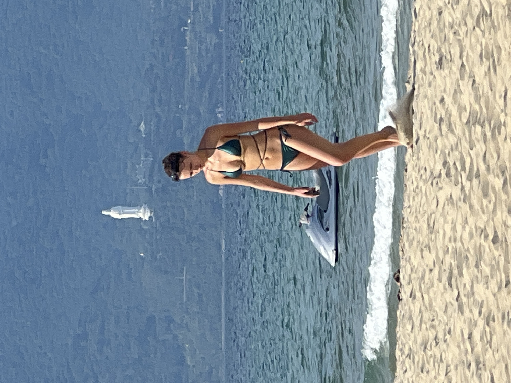 A woman in a dark green bikini walks on a sandy beach. She is looking down and to her left. Her hair is dark and pulled back. Behind her, the sea is a vibrant blue and green. A white jet ski is partially submerged in the water. In the distance, a large white statue of a woman stands on a green, forested mountain. The sky is a hazy blue. The scene appears to be in Da Nang, Vietnam, likely near the Marble Mountains where such a statue is located.