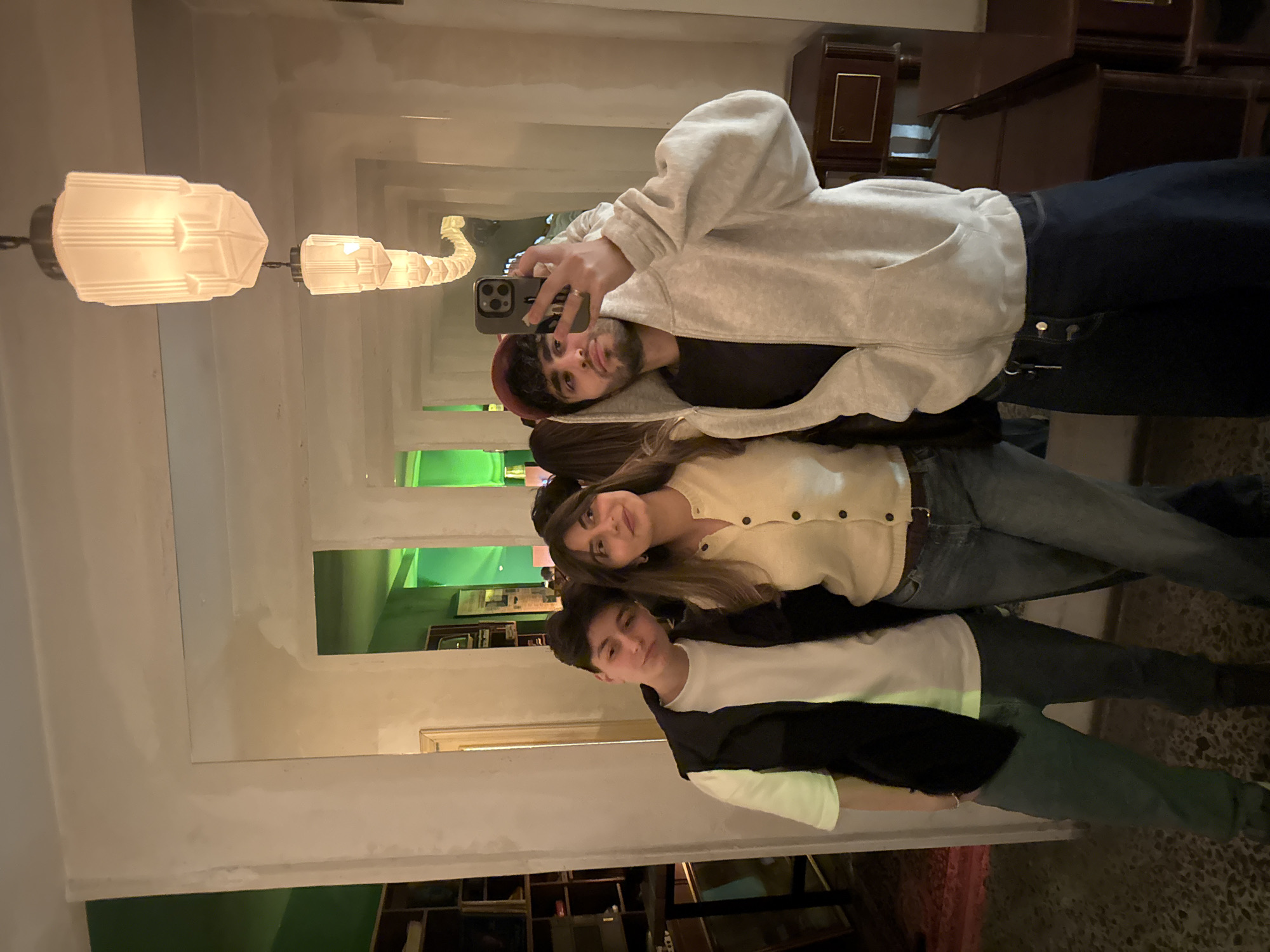 A group of three young people is posing for a selfie in what appears to be Yerevan, Armenia, as can be discerned from architectural and interior design elements. The person taking the photo is in the top position, wearing a light grey hoodie over a black shirt, and a red cap. They are holding a smartphone in their right hand. The person in the middle has medium-length brown hair and is wearing a light yellow cardigan with buttons. The person in the bottom position is wearing a white t-shirt under a black jacket. The three individuals are stacked close together in what may be an establishment featuring shelves. The background is blurred, but architectural and interior design elements can be seen, including light fixtures and decorative elements.