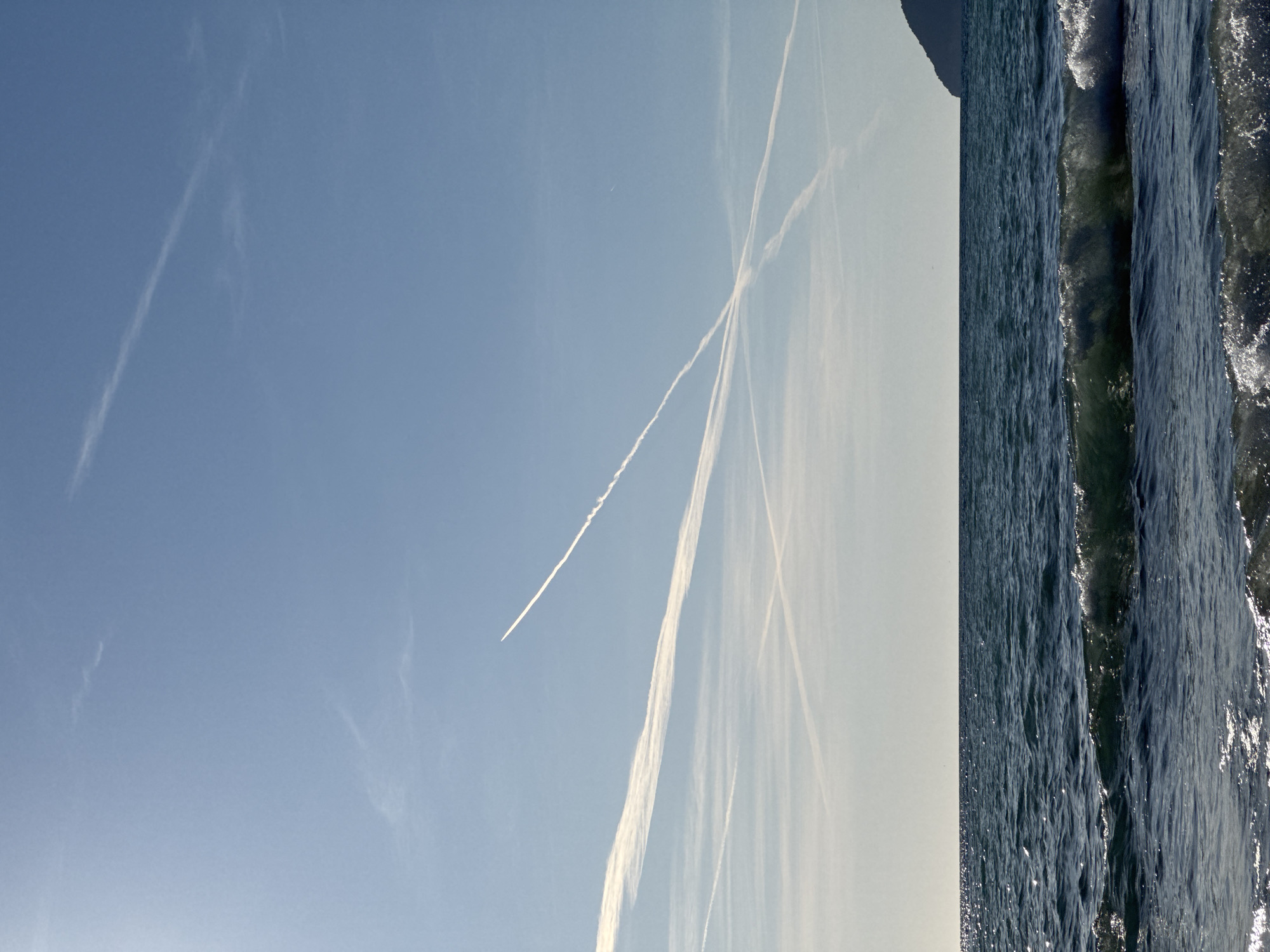 This image presents a broad outdoor scene, composed primarily of a clear sky and a body of water, consistent with the coastal environment of Bar, Montenegro.
The upper and left portions of the frame are dominated by a vast, vibrant blue sky. Several thin, white contrails from aircraft are visible, crisscrossing the azure expanse. One particularly prominent and fresh contrail stretches diagonally across the sky, with what appears to be a minuscule aircraft at its origin point, though the plane itself is barely discernible. The sky exhibits a subtle gradient, appearing a deeper blue overhead and transitioning to a lighter, somewhat hazy shade towards the horizon line.
The right side and lower portion of the image feature a deep blue body of water, likely the Adriatic Sea. The surface is characterized by active waves, with their crests catching and reflecting bright sunlight, creating sparkling highlights across the water. On the distant horizon, a dark, indistinct landmass or mountainous outline is visible, partially shrouded in atmospheric haze, reinforcing the Montenegrin coastal setting.
The scene depicts a bright, clear daytime, probably mid-morning or afternoon, given the intensity of the light and the high sun angle reflecting off the water. The weather appears fair and clear, allowing for the formation and persistence of the contrails in the upper atmosphere. No people, man-made objects other than the distant aircraft's contrails, or visible text are present in the image. The overall impression is one of natural grandeur and a peaceful, expansive environment.
