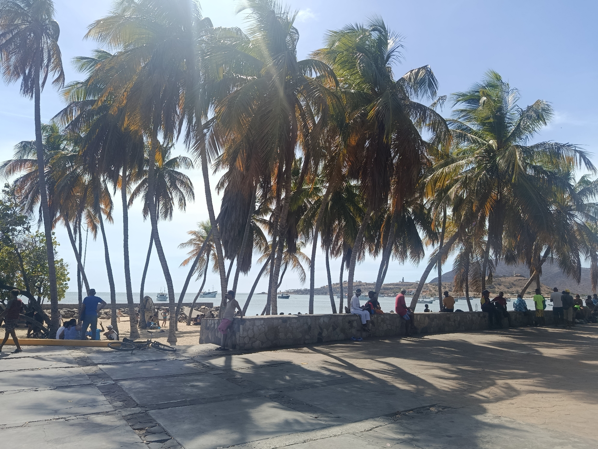 The image depicts a waterfront scene in Juan Griego, Venezuela. Palm trees line the shore, casting shadows on a paved area in the foreground. People are gathered in various groups along the beach and sitting on a low stone wall separating the pavement from the sand. In the background, boats are anchored in the bay, and a hillside with a lighthouse is visible. The sky is clear and blue.