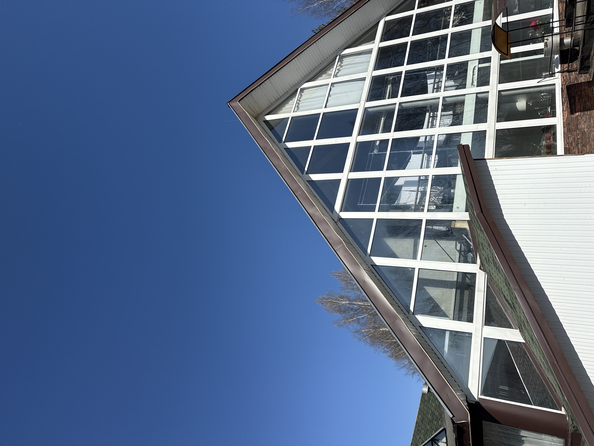 A low-angle shot shows a building facade against a clear blue sky. The building has a large, triangular window section with a grid of white frames. A balcony with a chair and some plants can be seen through the windows. The facade appears to be partly brick and partly white siding. Some bare tree branches are visible in the upper left of the frame. Given the prompt context, this image may be taken in Галево, Russia.