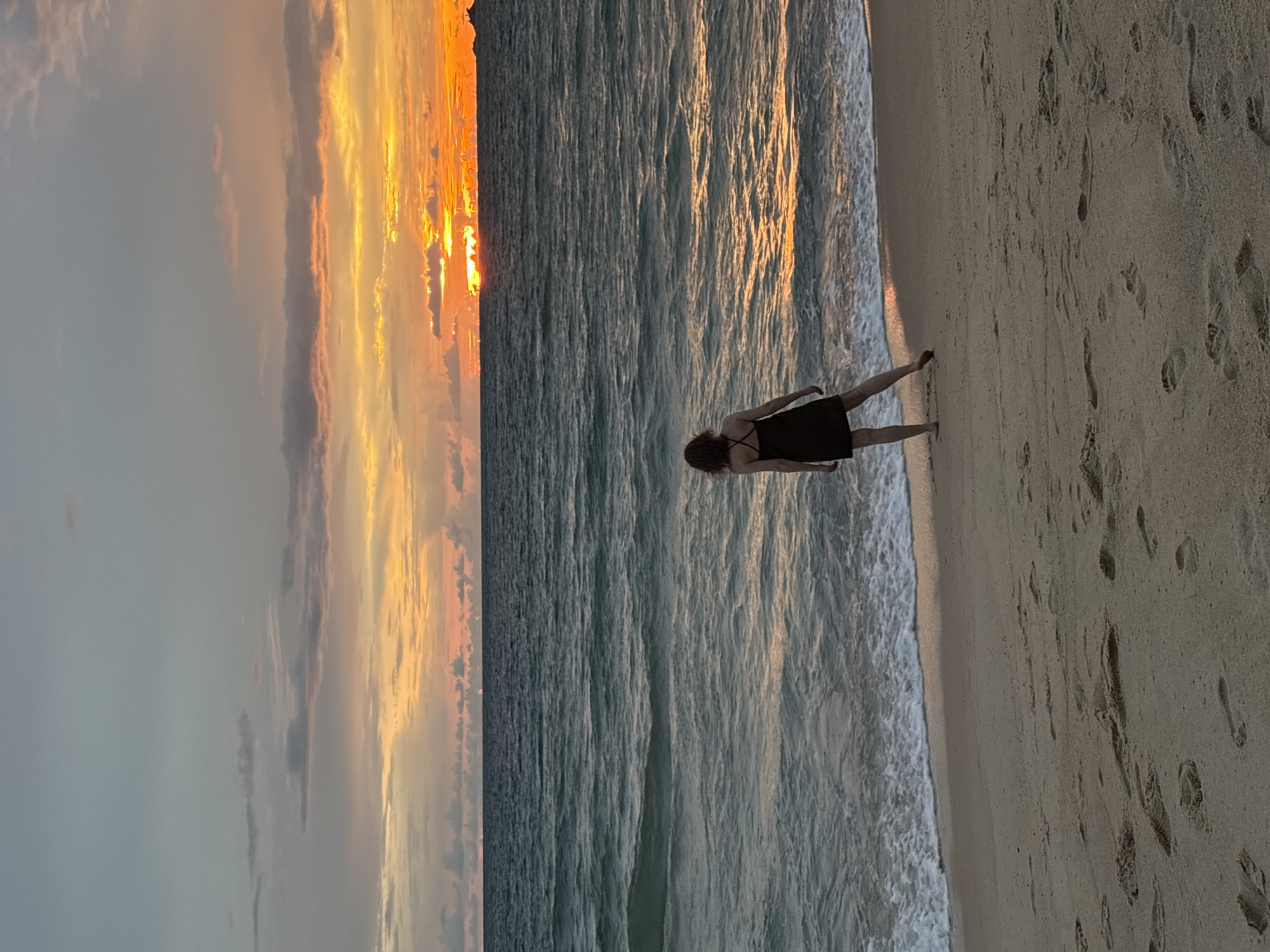 A woman stands on a sandy beach at sunset, facing the ocean. The sky is a mix of soft gray clouds and vibrant orange and yellow hues from the setting sun. The ocean waves are gently rolling onto the shore, creating a foamy white line where they meet the sand. Footprints are visible in the sand, indicating previous activity on the beach. The woman is wearing a dark-colored dress and appears to be looking out at the sunset. The overall mood is peaceful and serene. The time of day is sunset, and the weather appears calm. The location is a beach in Choeng Thale, Thailand. There is no visible text in the image.