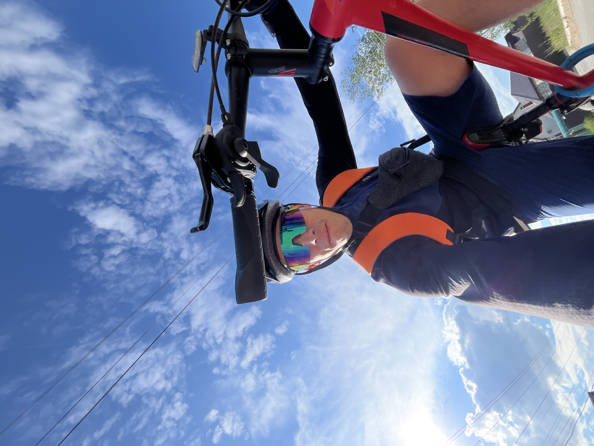 A low-angle, wide-shot perspective shows a cyclist on a red bicycle against a bright blue sky with scattered white clouds. The cyclist, wearing a black long-sleeved shirt, dark blue shorts, a black helmet, and rainbow-lensed cycling glasses, is looking towards the camera. An orange strap is visible over the cyclist's left shoulder, and a small black bag is attached to their waist. The bicycle's handlebars, stem with "DIRECT TECH" branding, and part of the frame are prominent. Power lines run diagonally across the upper portion of the frame. In the background, a few houses with brown roofs and green foliage are partially visible. The sun is positioned on the left side of the image, creating lens flare and illuminating the clouds. The scene appears to be in a suburban or rural area, possibly on a street in Chaikovsky, Russia, given the presence of houses and overhead power lines under a clear sky.