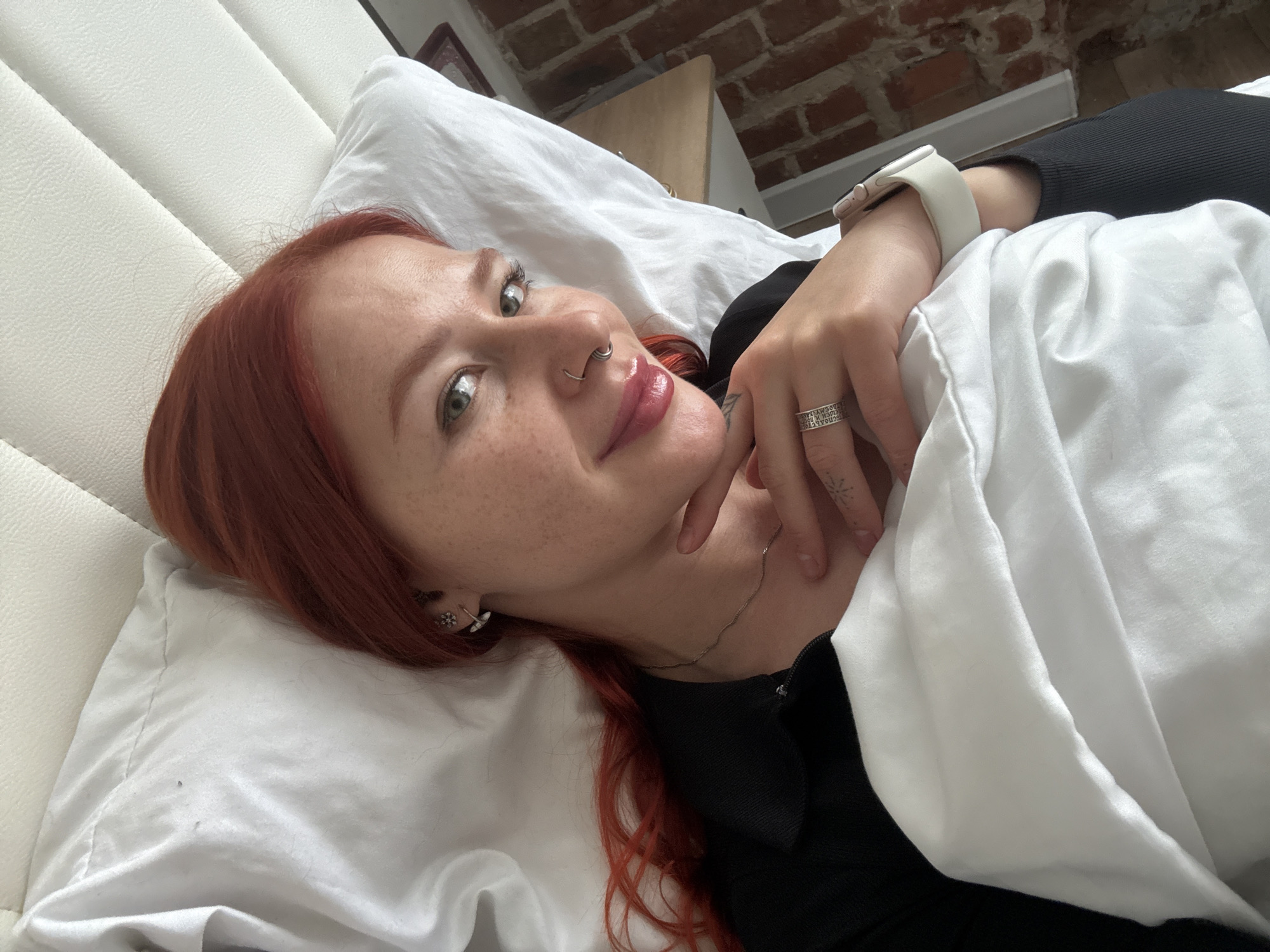 A woman with red hair is lying on a white bed. She has a nose ring and freckles. She is wearing a black top and a white watch. Her hand is resting on her neck, revealing a ring and a small tattoo. The background includes a white headboard and a brick wall. There is no indication of the location being Rostov-on-Don, Russia.