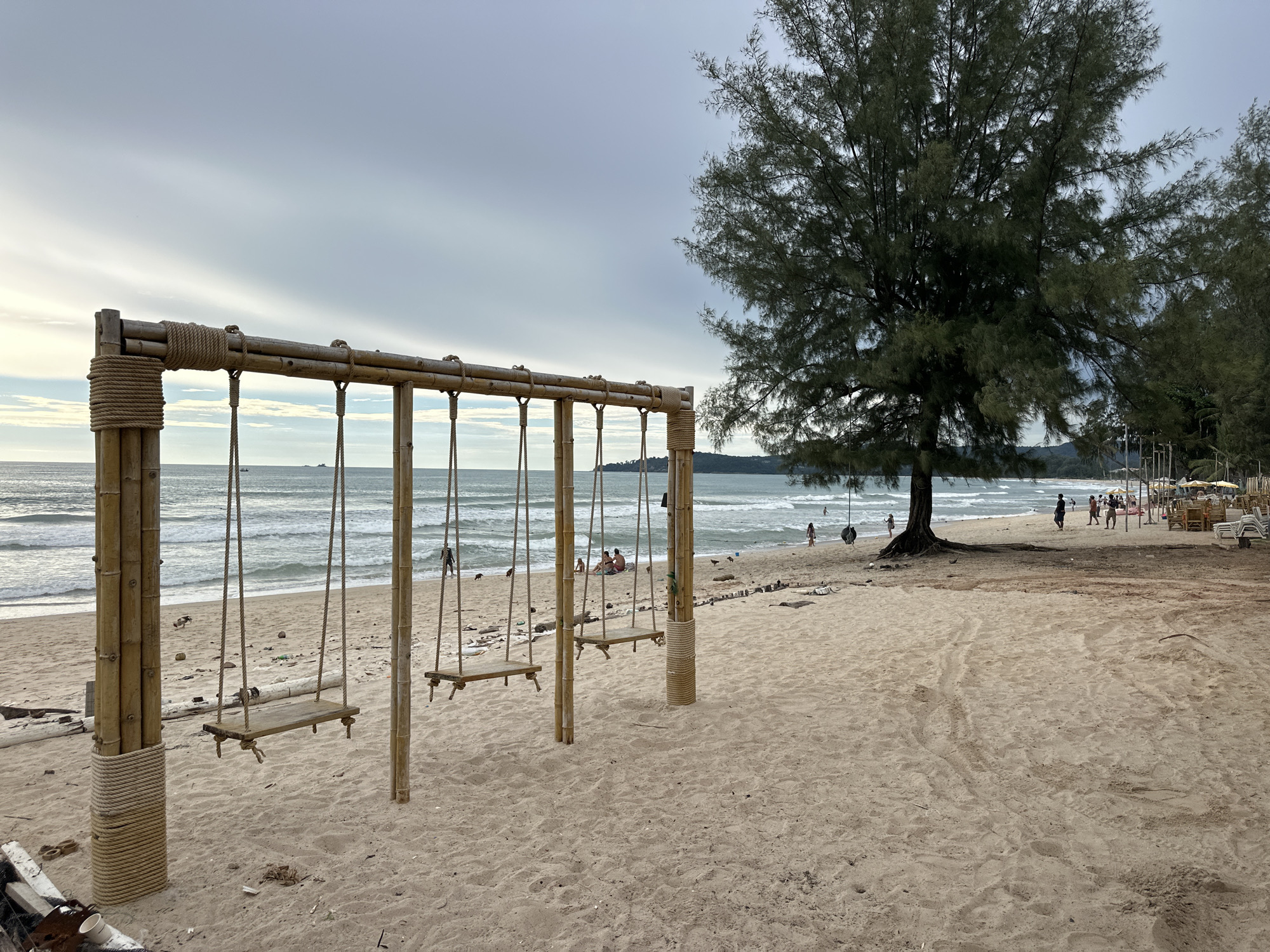 A wide shot captures a tranquil beach scene in Choeng Thale, Thailand, under an overcast sky. In the foreground, a bamboo swing set with four seats rests on the sandy shore, its natural materials blending with the environment. The ocean, with its gentle waves, stretches towards the horizon, where a few boats are visible. A large, mature tree stands prominently on the right, its branches reaching out over the beach. Several people are scattered across the sand, some walking, others relaxing near the water, and a group is seated together on the sand. The beach is dotted with debris, including driftwood and scattered items, suggesting a natural, unmanicured setting. In the distance, a row of beach chairs and umbrellas indicates a resort or recreational area. The overall atmosphere is one of peaceful leisure.