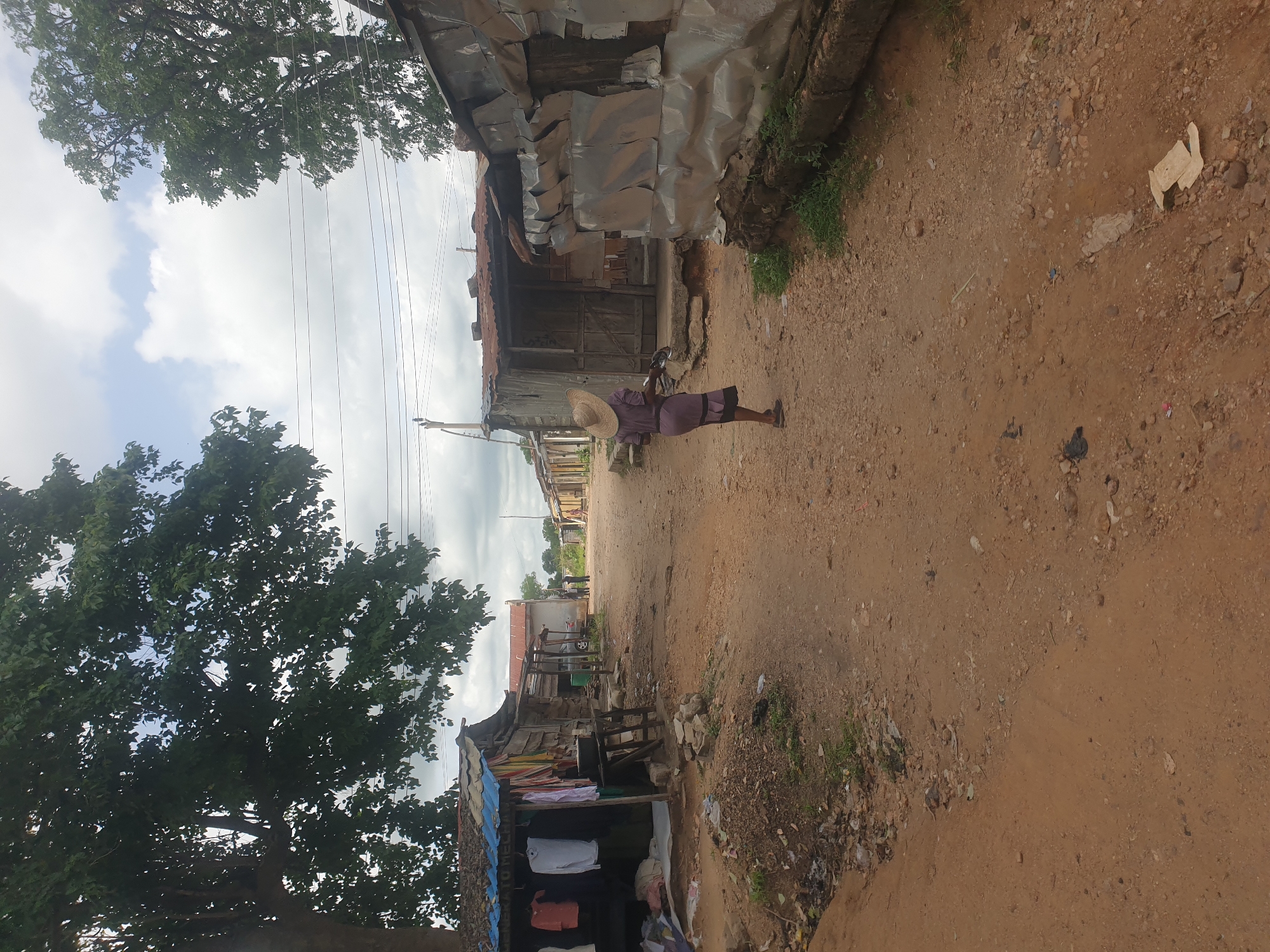 A vertical outdoor shot depicts a dusty dirt road lined with shacks in Unknown, Unknown. A woman wearing a purple dress and a straw hat walks along the road, pushing a bicycle. To the left and right of the road, shacks are visible. Some appear to be made of corrugated metal, while others are constructed from wood and covered with tarps. In the lower left, clothes hang from a shack, seemingly for sale. Overhead, power lines stretch across the sky, framed by trees. The sky is overcast with a mix of clouds. The road is made of dirt and appears to be uneven with scattered debris.