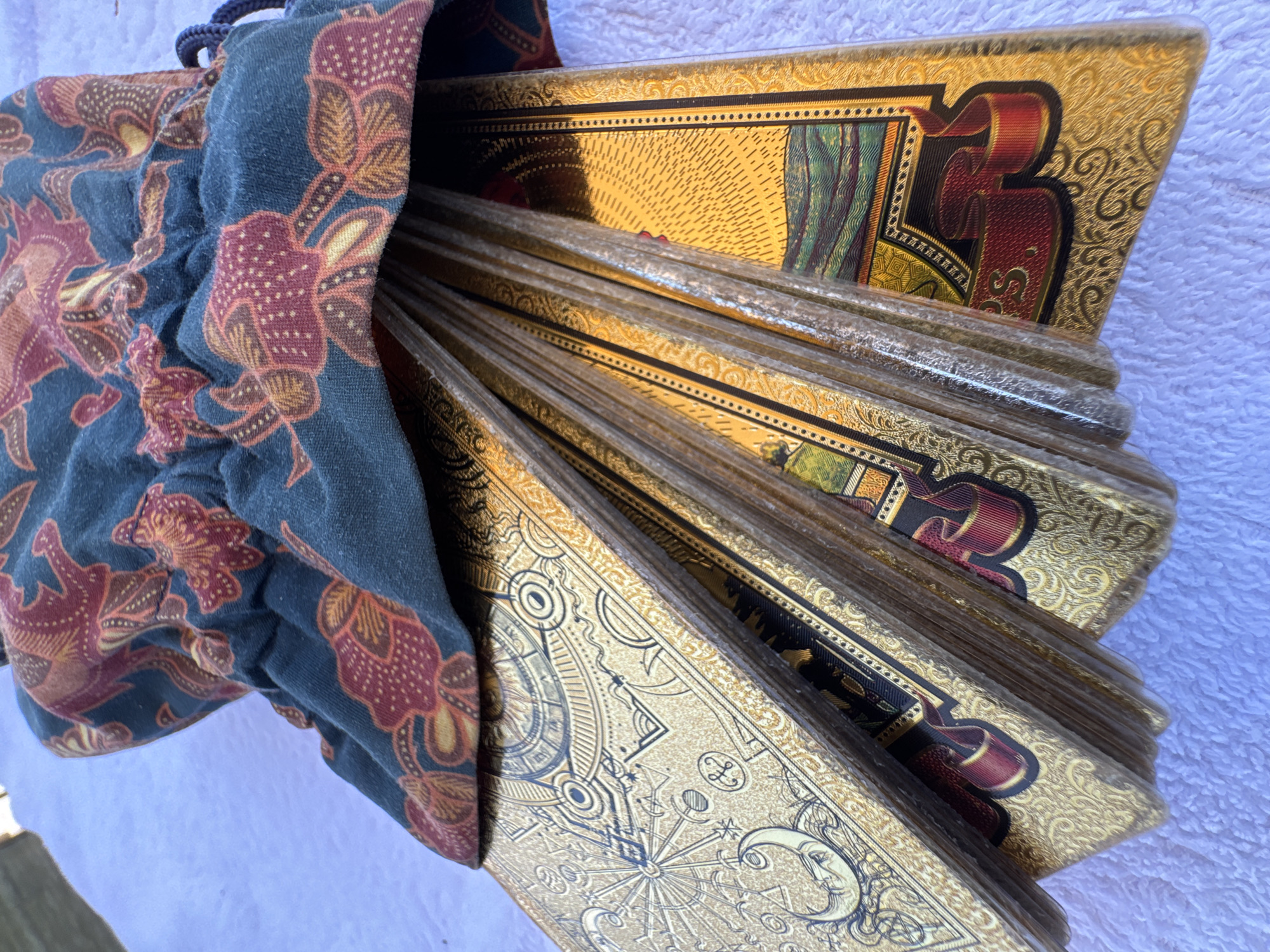 A close-up shot shows a pile of gold-edged tarot cards protruding from a blue drawstring bag with a red and brown floral pattern. The cards display intricate designs with astronomical symbols and ornate patterns. The bag is situated on a textured white surface. There is no visible evidence to suggest that this image was taken in Đà Nẵng, Vietnam. There are no people visible.