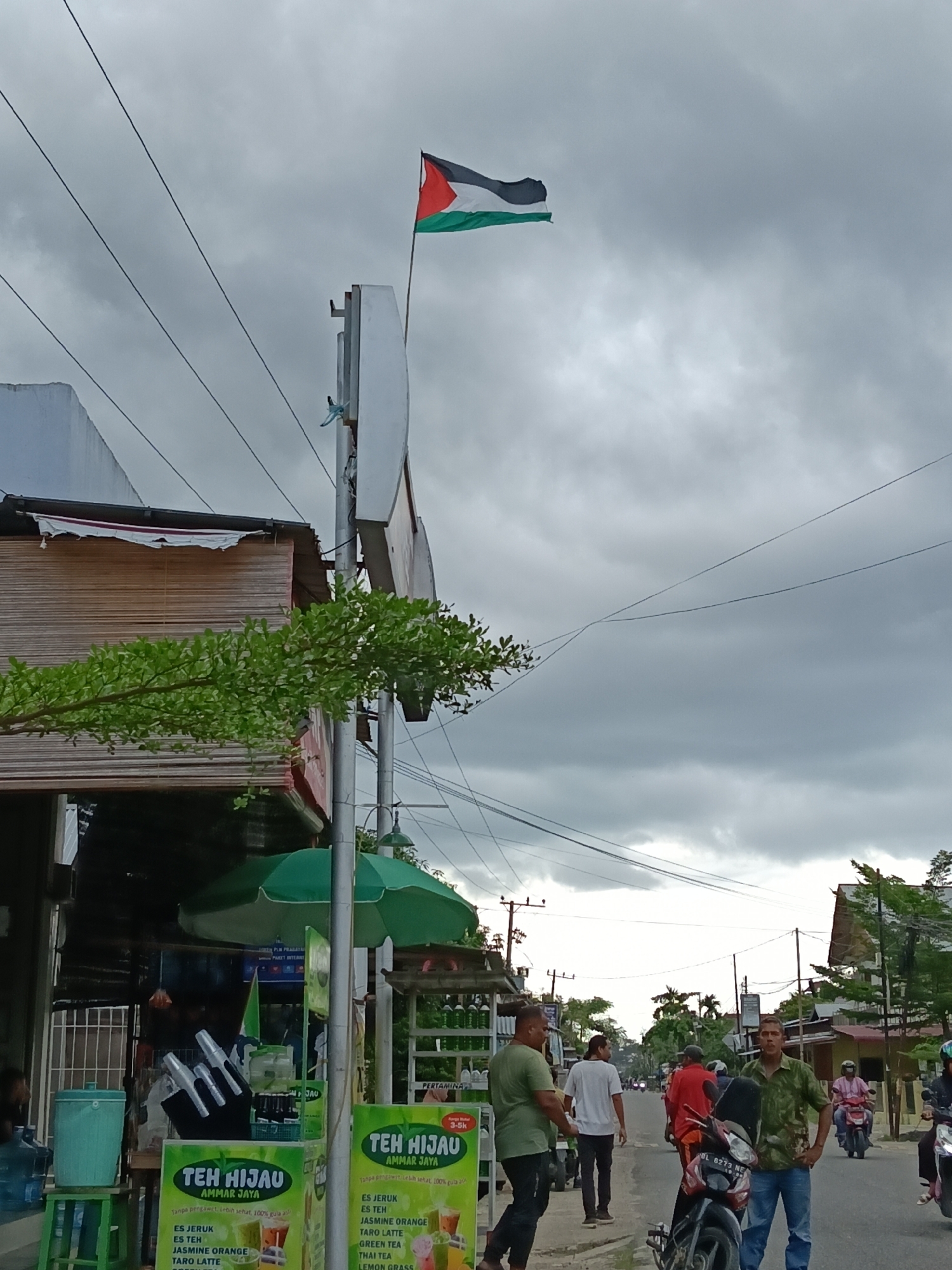 Here is a detailed description of the image:
**Scene Overview:**
The image captures a street scene in what appears to be a residential or commercial area in Lhokseumawe, Indonesia. The sky is overcast, suggesting it may be a cloudy day.
**Subjects and People:**
Several people are visible in the image.
* There are several men standing on the side of the road.
* Other people are seen in the background, some of whom are riding motorcycles.
**Objects:**
* A Palestinian flag is prominently displayed atop a tall pole.
* A small food stall or vendor stand is selling "Teh Hijau" (Green Tea), advertising various flavors like jasmine orange and taro latte.
* Several motorcycles parked or driving along the street
* Electricity poles and wires are present in the background.
**Actions and Interactions:**
* People are walking along the street and seemingly conducting everyday activities.
* The stall vendor appears to be preparing drinks.
**Environment:**
* The scene is on a paved street in an urban area.
* Buildings are visible on both sides of the street, suggesting a mix of residential and commercial establishments.
* The foliage and trees hint at a tropical environment.