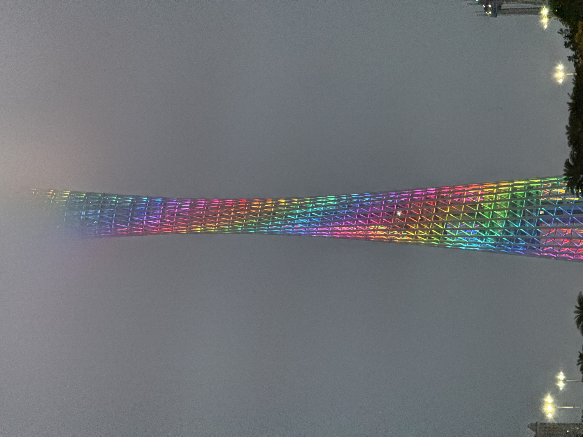 The image shows the Canton Tower in Tianhe District, Guangzhou, China, illuminated with rainbow colors. The sky is overcast and hazy, obscuring the top and bottom of the tower. The lower portion of the image has lights and trees. The photo is taken from an elevated position. The building is lit up with colors like red, orange, yellow, green, blue, indigo, and violet.